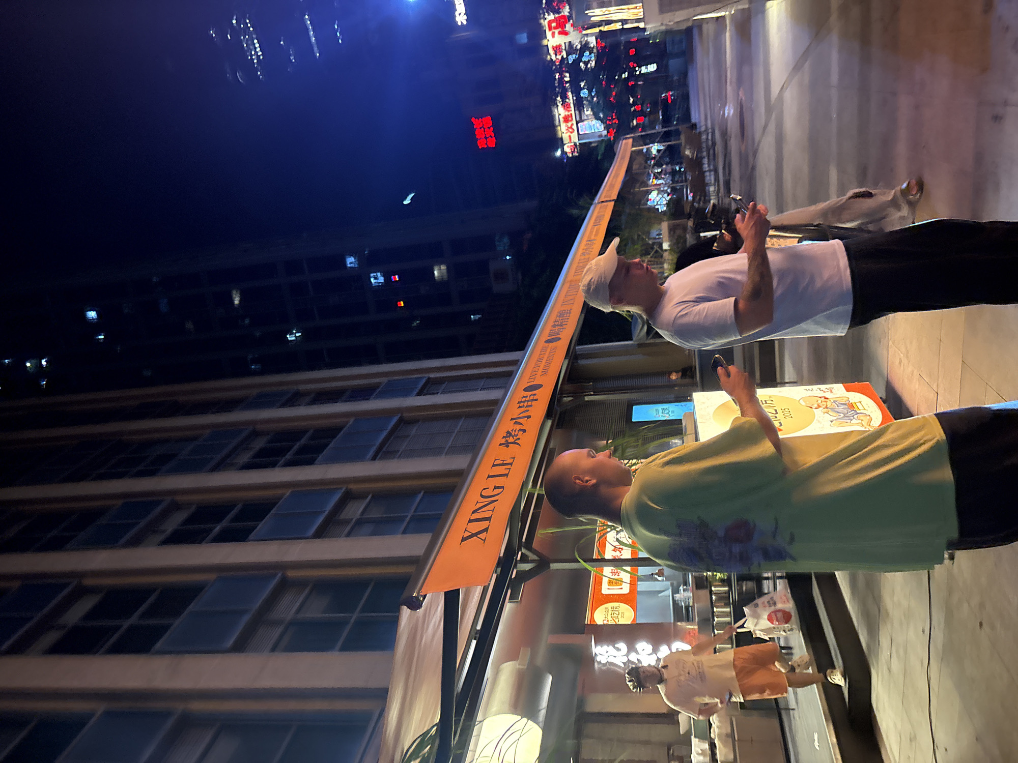 In Haizhu District, China, at nighttime, two young men are standing on a sidewalk in front of a restaurant. The man in the foreground, wearing a light green t-shirt and dark pants, is facing away from the camera and looking down at his phone. To his left, a man in a white t-shirt and a white baseball cap is also looking at his phone. Behind them, the restaurant has an orange awning with Chinese and English text that reads "XING LE" and "烤小串". Another man in an orange t-shirt and shorts is walking up stairs to the left of the restaurant carrying a white bag. The background shows tall buildings with illuminated windows, a dark night sky, and streetlights reflecting on the wet pavement.