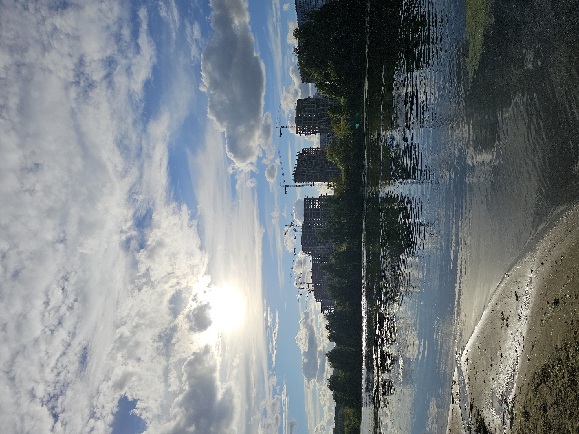 A wide shot captures a body of water with a sandy shore in the foreground, leading to a dark line of trees. Behind the trees, an urban skyline features numerous high-rise buildings under construction, with several cranes visible. The sky above is a mix of blue with scattered white clouds, and the sun is partially obscured by clouds, casting a bright glow. The water reflects the sky and the distant buildings.