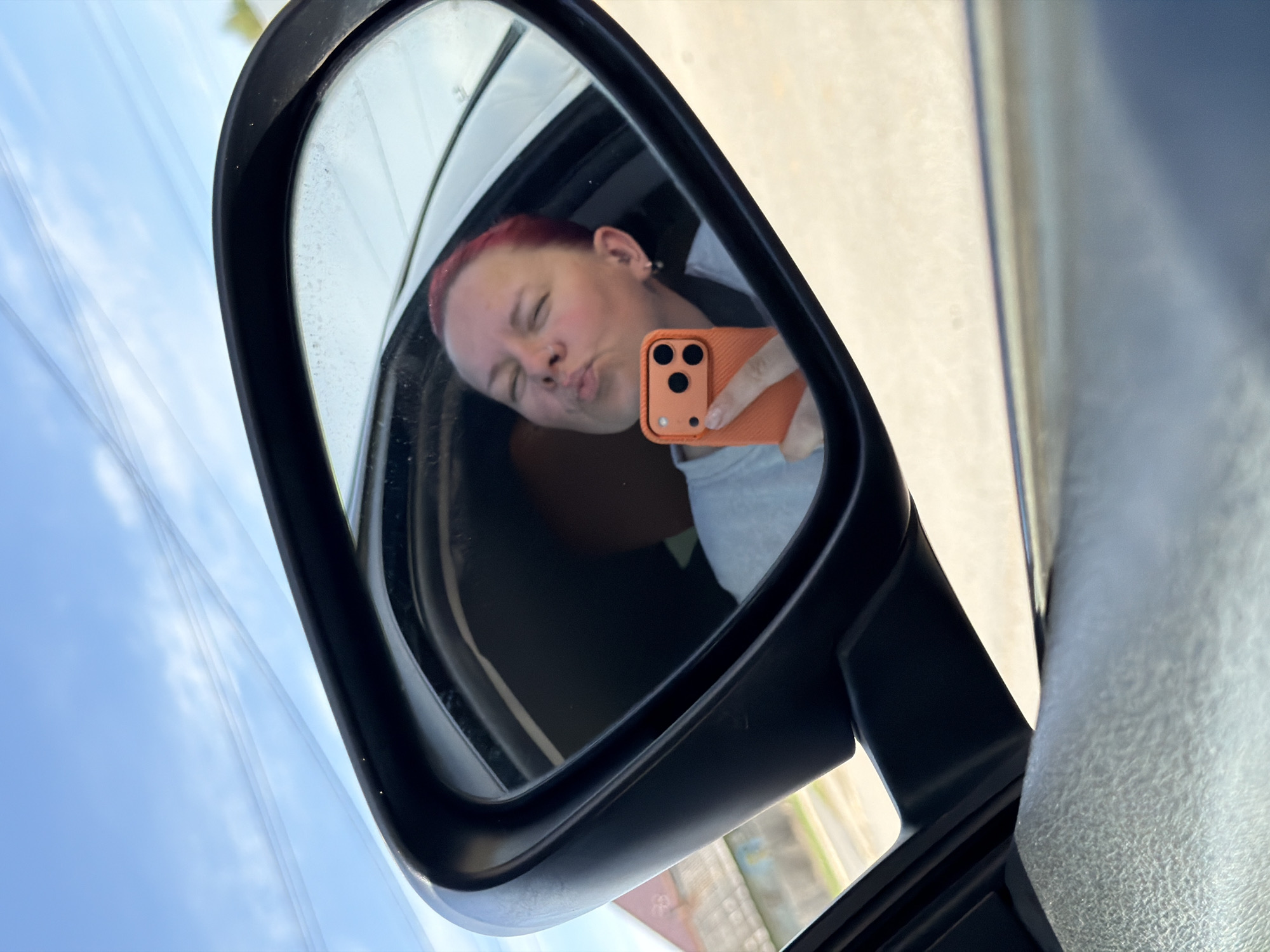 A reflection in a car's side mirror shows a person with short red hair and fair skin holding an orange smartphone up to their face, pursing their lips in a kissy face expression. The person is wearing a light grey or blue shirt. The car's interior is partially visible. The background seen in the mirror suggests an outdoor setting with buildings and greenery, possibly an urban or suburban area. Outside the car window, power lines are visible against a light blue sky. The location is presumably Chaikovsky, Russia, though no explicit details in the image confirm this.