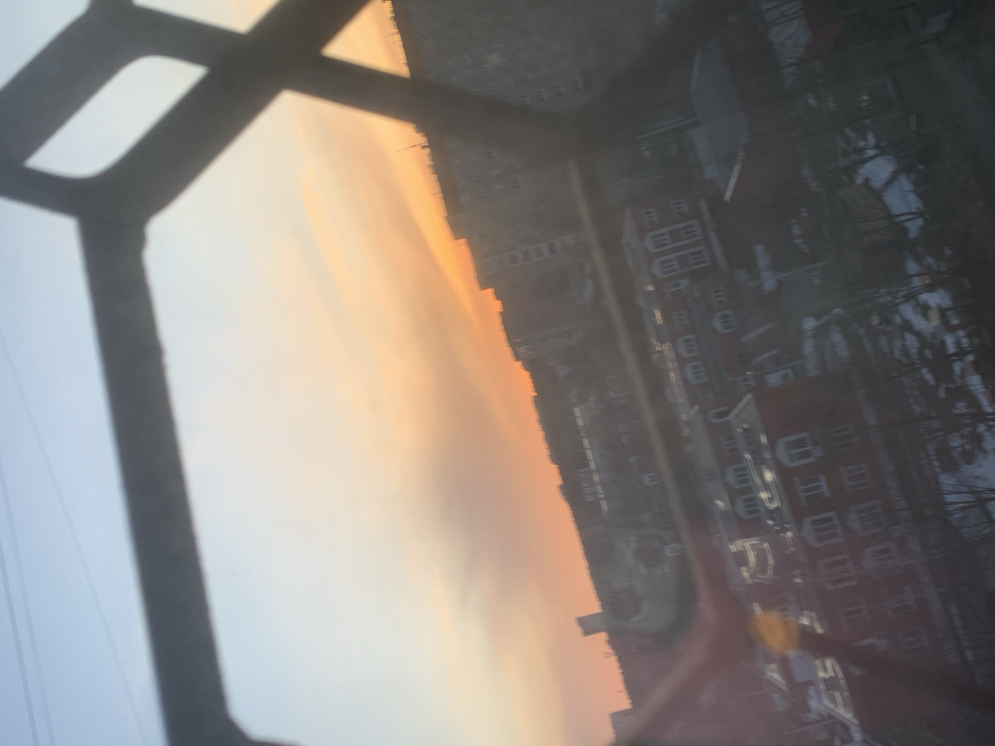 The image shows a view from a window with a metal frame. Outside, a cityscape is visible during what appears to be sunset or sunrise, given the orange-colored sky. Buildings with multiple windows are visible, as well as bare trees, possibly indicating winter or early spring. While the exact location in Izhevsk cannot be determined from this image, the architectural style of the buildings suggests an urban setting.
