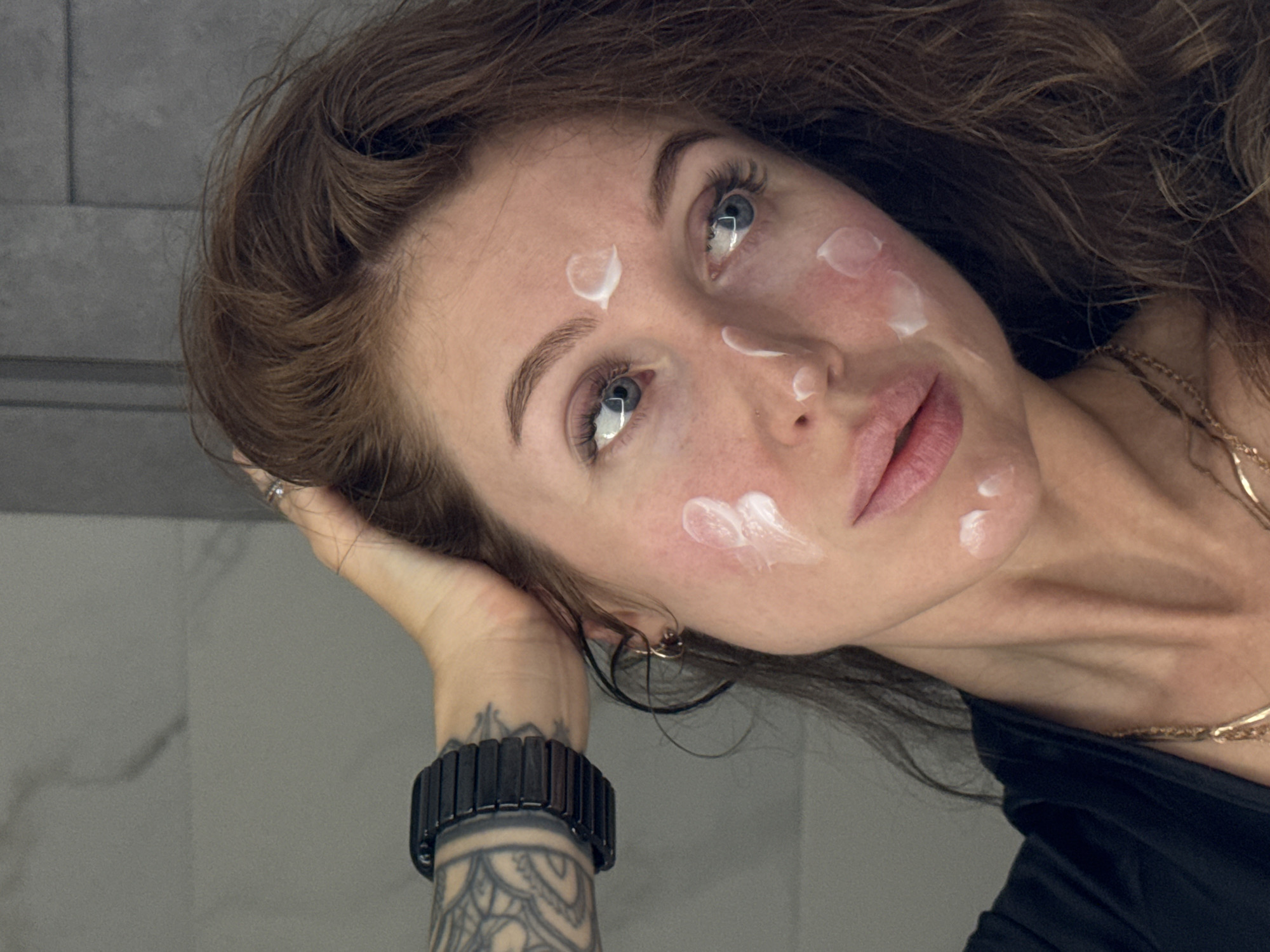 A woman with auburn hair is lying down, her head tilted to the side, with a neutral expression on her face. Her skin is fair with a few dots of white cream on her forehead and cheeks. She has blue eyes, well-defined eyebrows, and pink lips. Her hand is raised near her head, and she is wearing a black bracelet on her wrist, which features a tattoo of a circular design. She is wearing a black top and a gold necklace. The background is a gray, textured wall, likely of a bathroom. The image seems to be taken indoors. There is no definitive evidence that this image was taken in Chaikovsky, Russia.