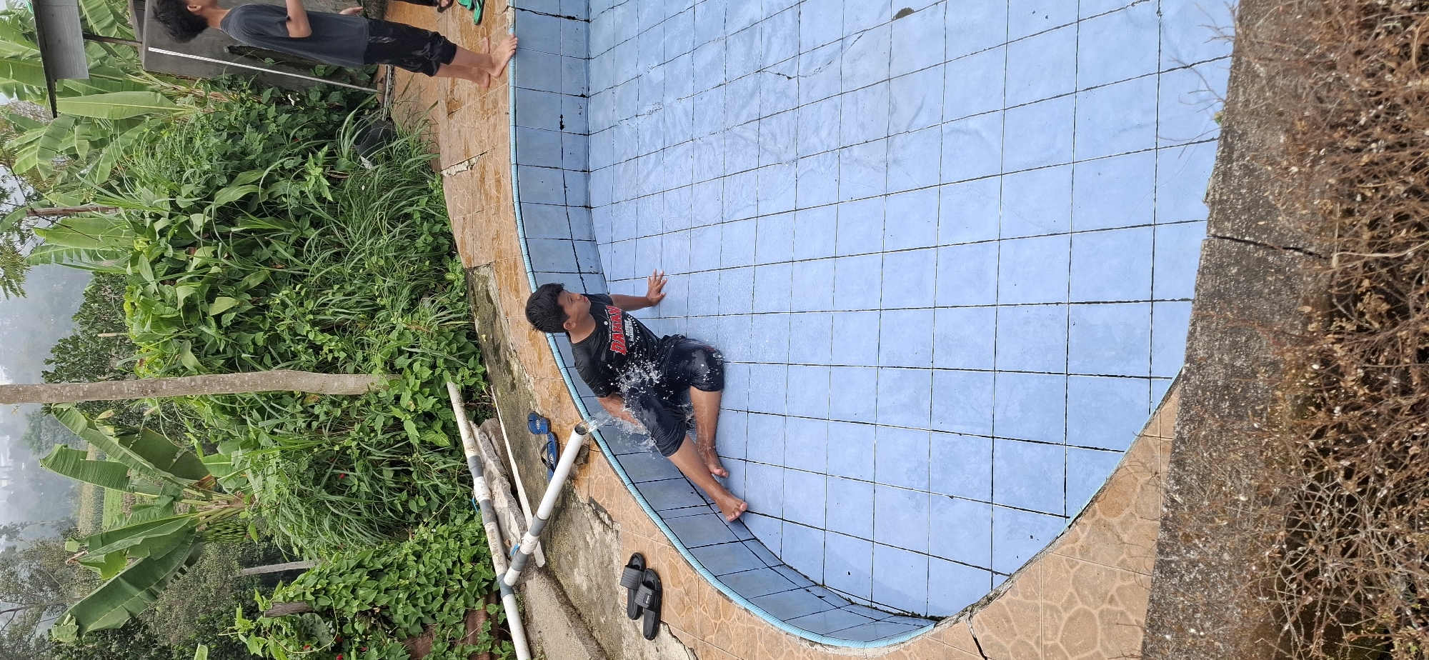 The image depicts an outdoor scene with a young person sitting in an empty, tiled swimming pool. The person, wearing a black t-shirt with red writing and dark shorts, is splashing water, creating a spray around them. The pool has blue tiles and a tan tile border. Outside the pool, there are several pairs of flip-flops scattered on the ground. In the background, lush green vegetation, including banana trees, is visible. A pipe system is also present near the edge of the pool, with water flowing from it into the pool. The overall environment suggests a rural or semi-rural setting, potentially in Puntukrejo, Indonesia, given the presence of tropical foliage. Another person, partially visible in the upper right of the frame, stands outside the pool, also wearing dark clothing.
