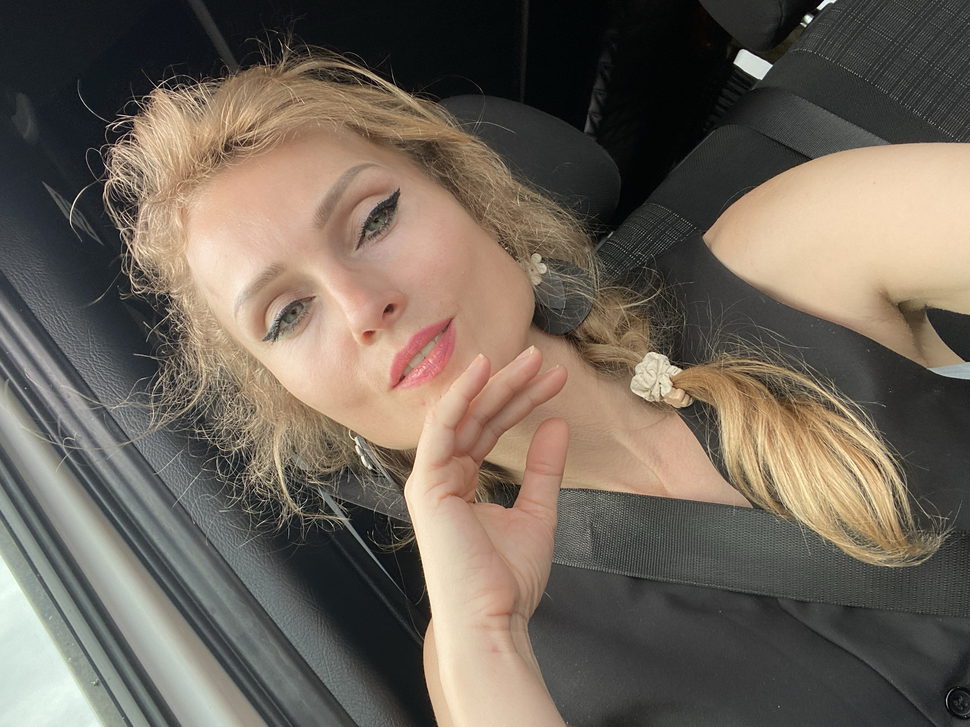 A blonde woman with green eyes and pink lipstick poses for a selfie in a car. She is wearing a black vest and a seatbelt. Her hair is pulled back in a braid with a scrunchie. The background of the image shows the interior of a car, with a black leather seat and a window. The image was taken in the city of Валожынскі сельскі Савет, Belarus.