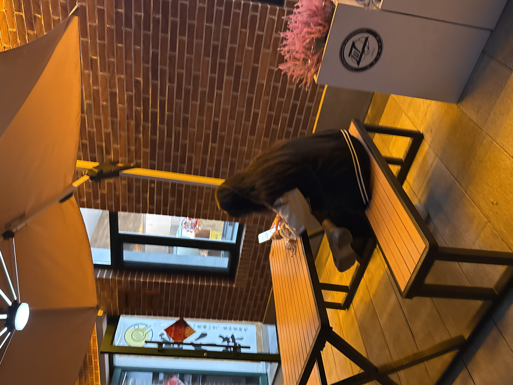 A person wearing a black jacket and gray pants is sitting at a table with a phone in their hand. The table is made of wood and metal. There's an umbrella above. The photo has been taken from a low angle and is slightly rotated. The background shows a brick wall, a window, and signs in Chinese. A flower box with pink flowers can be seen on the right. The location is likely Zhongtun, China based on the prompts.