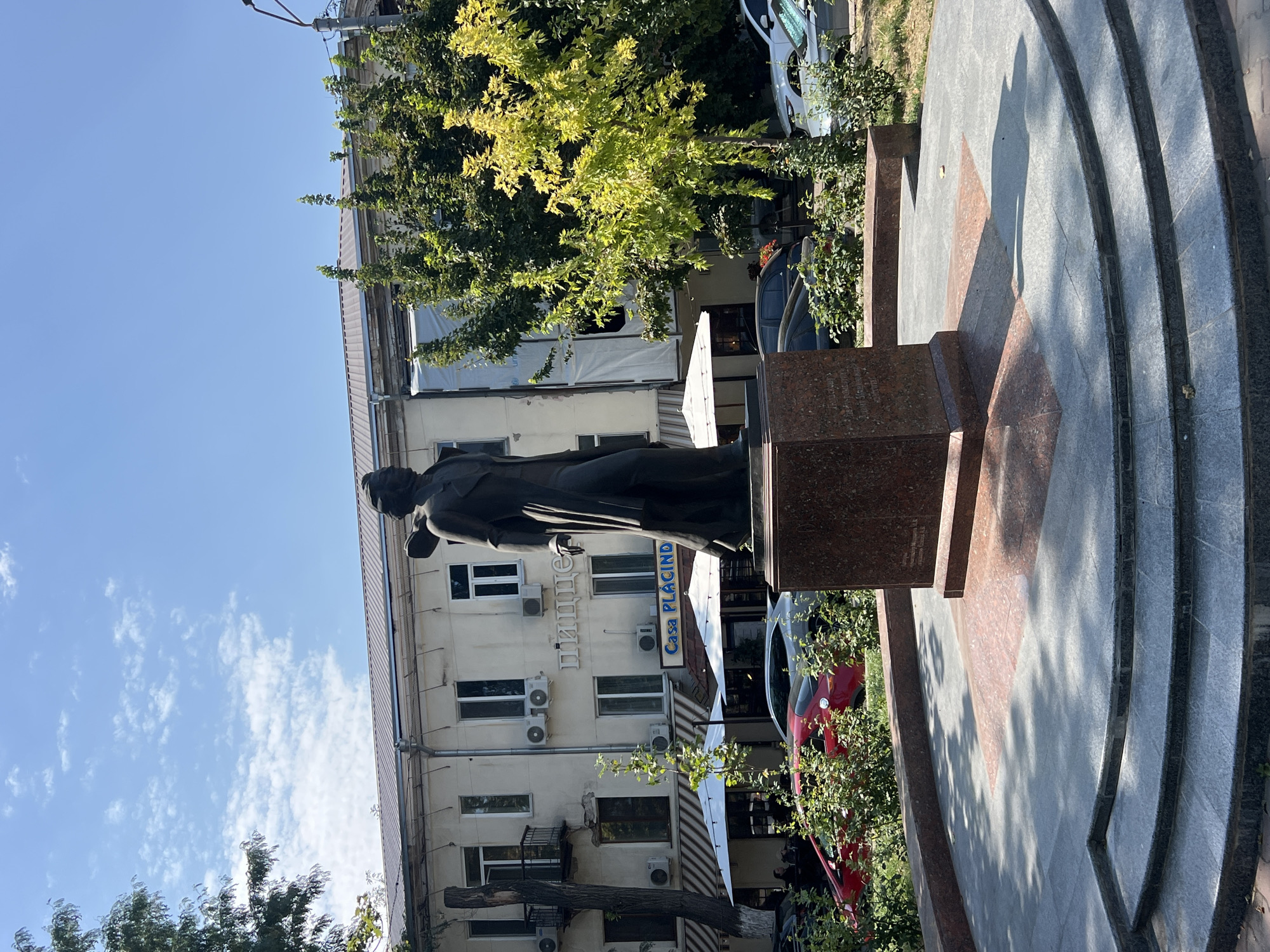 In Odesa, Ukraine, a bronze statue of a historical figure stands in a city plaza. The statue depicts a man in period clothing, facing right, with his left hand extended forward. The statue is mounted on a granite pedestal. Behind the statue, a light-colored building with multiple windows and air conditioning units is visible. The building has awnings over some of the ground-floor windows and signage that reads "Casa PLACIDO." To the right of the building, lush green trees with bright yellow leaves are in full bloom. In the foreground, the plaza is paved with gray and red stone tiles, forming a circular pattern around the monument. Several cars are parked in the background, and greenery lines the perimeter of the plaza. The sky is a clear blue with scattered white clouds.