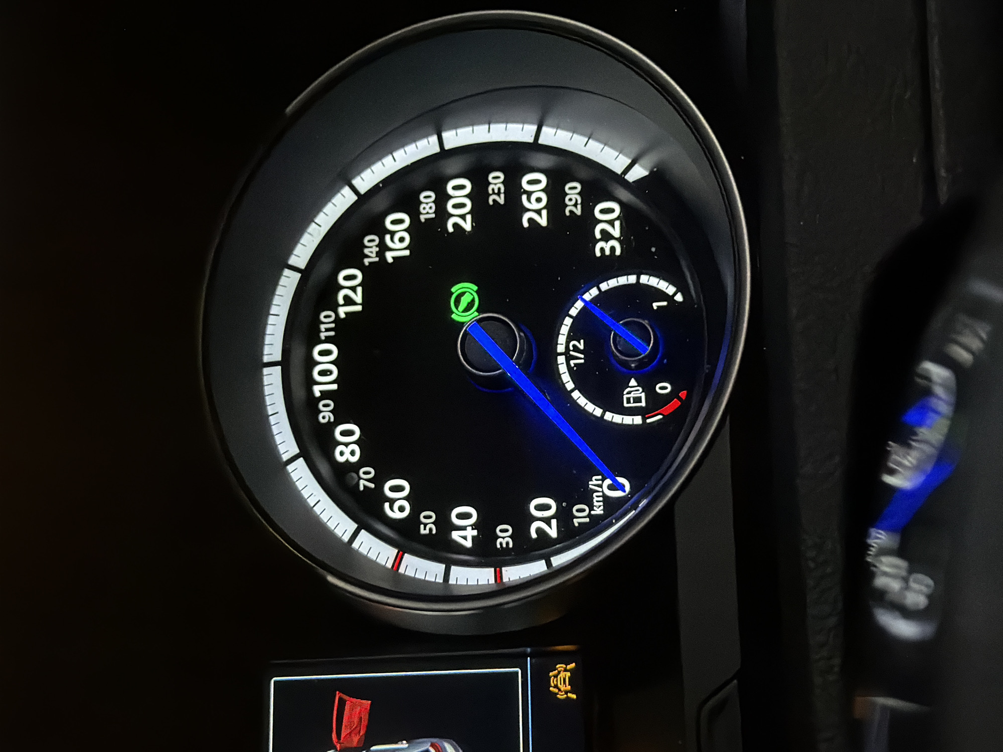 The image shows a close-up view of a car's dashboard, focusing on the speedometer and other indicators. The speedometer displays speeds in kilometers per hour, with markings from 0 to 320. The current speed indicated by the needle is approximately 45 km/h. A fuel gauge is also visible, showing the fuel level slightly above the empty mark. To the left of the speedometer, a small digital display shows a red icon that appears to be a shopping cart with a flag, and below it, a yellow icon resembling a car with sensor waves emanating from it. The background is dark, suggesting the interior of a vehicle at night or in a dimly lit environment. There are no people visible in the image. The image was likely taken inside a car in Sofia, Bulgaria, given the context of the request.