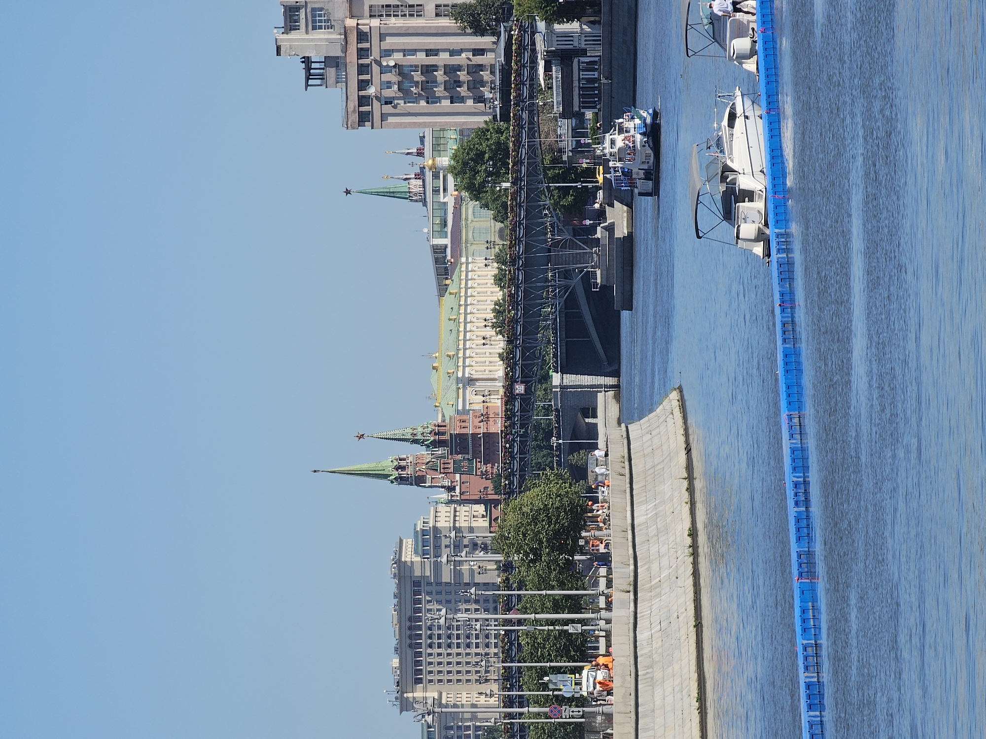The image shows a body of water with several boats on it, as well as a cityscape in the background. The water appears to be a river, and there is a long blue floating barrier in the foreground. On the water, there are two white motorboats, each with an awning and several people on board. In the distance, a larger white and blue passenger boat is visible, with more people on its decks.
The cityscape includes several buildings, including what appear to be historical structures with green spires and red brick walls, likely part of the Kremlin. A bridge with metal supports spans the water in front of these buildings. The sky is clear and blue, indicating a sunny day. On the bank in the middle ground, there are trees, lampposts, and several people, some of whom are wearing orange vests, suggesting construction or maintenance work. The overall scene is one of a bustling urban environment on a clear day, with a focus on the water and the boats. The location is identified as Балтия Кп, Russia.