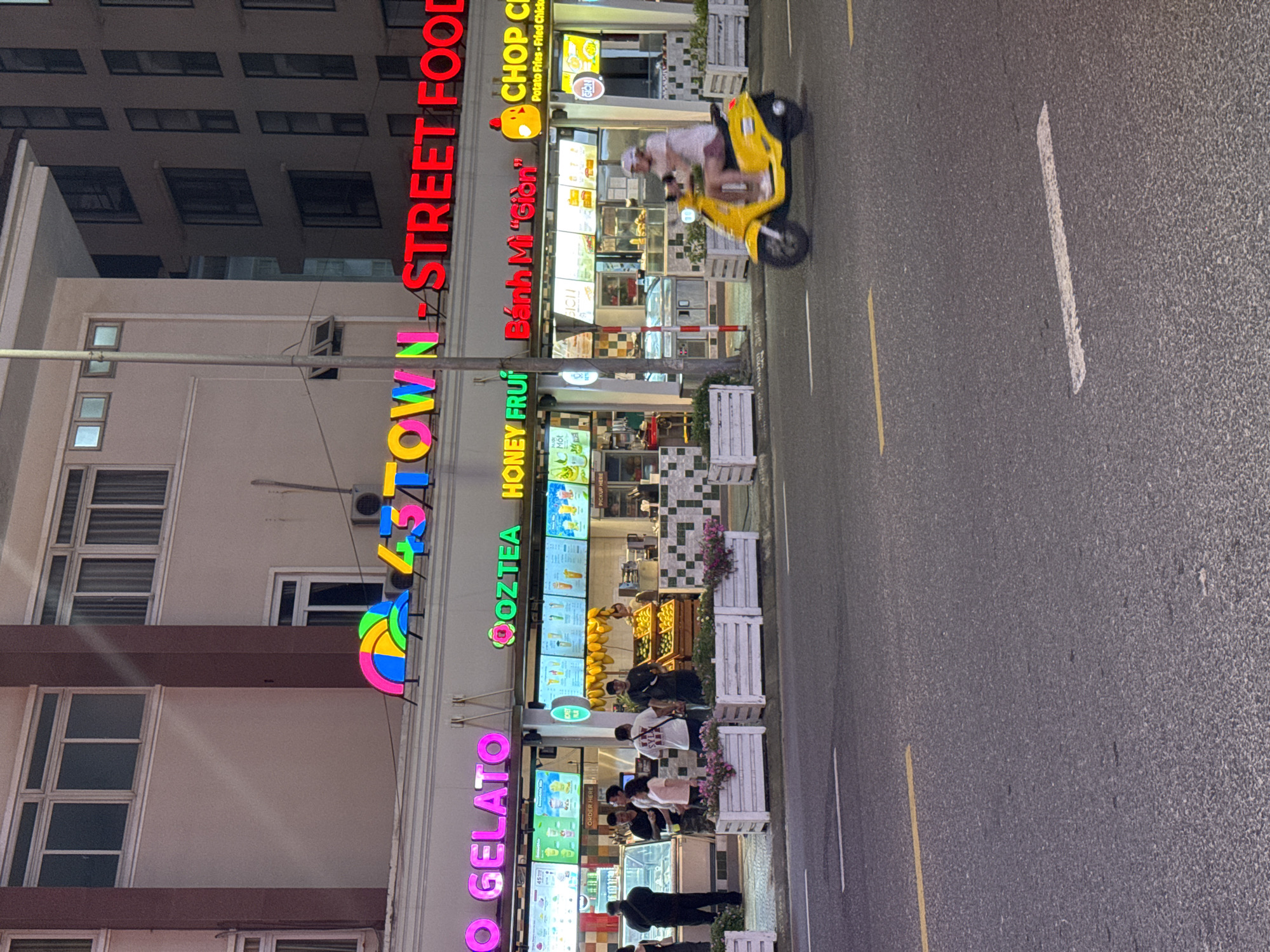 The image captures a vibrant street scene in Đà Nẵng, Vietnam, with the storefront of "O Gelato" prominently featured. The building's facade is adorned with colorful neon signs, including "O GELATO," "DA'NASTOWN STREET FOOD," "OOZTEA HONEY FRUIT," and "Bánh Mì Giòn." People are gathered outside the various food establishments, including "CHOP CI," interacting with the businesses. A person is riding a yellow scooter on the street. The street has yellow and white lane markings. The buildings are multi-storied with rectangular windows. The image has an evening aesthetic due to the lighting.