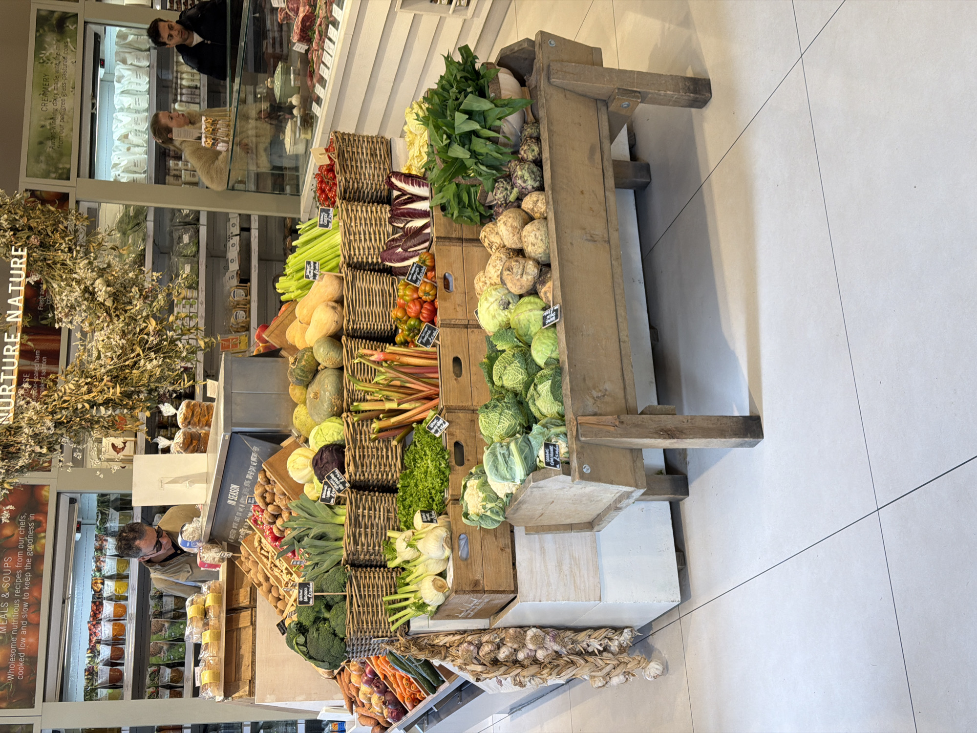 This image showcases a well-stocked produce display, possibly inside a grocery store or farmer's market. The location appears to be in the city of London, United Kingdom, indicated by "Creamery" information within the store.
**Subjects/Objects:**
* A large wooden produce display stand.
* Assortment of fresh vegetables and some fruits, including cabbages, squash, tomatoes, celery, garlic, and various leafy greens.
* "Meals & Soups" advertising display.
* "Creamery" details highlighting award-winning dairy.
**People:**
* A man looking at prepared "Meals & Soups" inside the store.
* Other possible customers or staff in the background near a refrigerated display.
**Environment:**
* Brightly lit indoor space with tiled flooring.
* Refrigerated displays and shelving visible.
**Actions/Interactions:**
* A customer is observed browsing prepared foods.
* Produce displayed for purchase.
The overall impression is of a high-quality grocery store or market that emphasizes fresh, local ingredients.