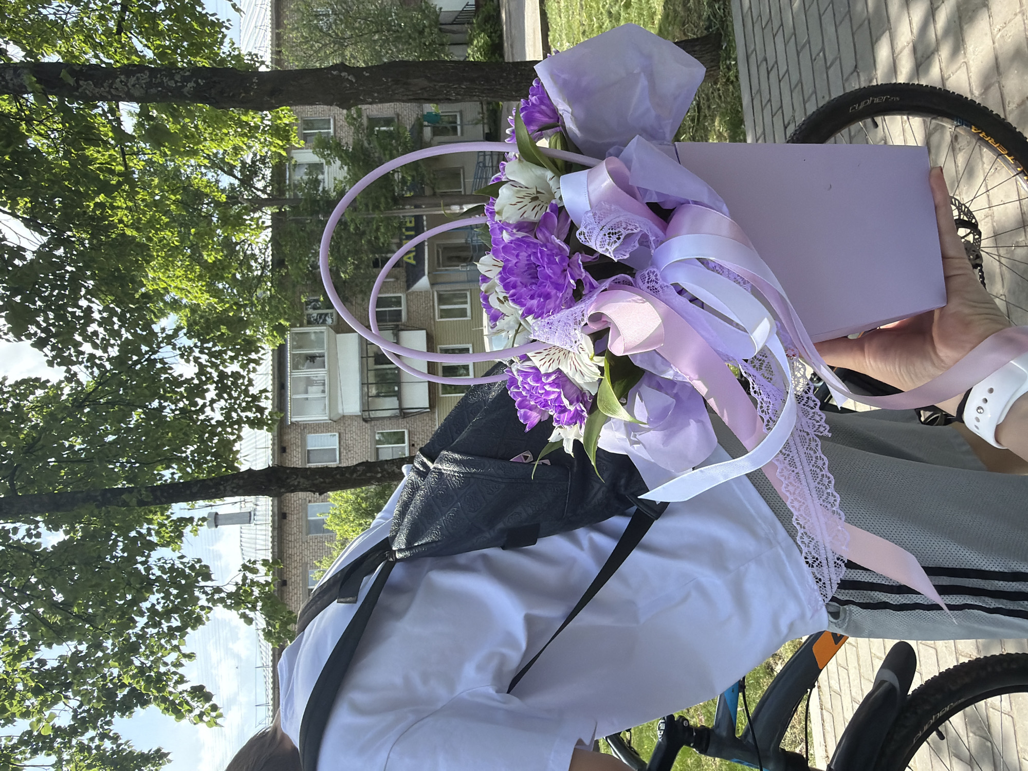 A person on a bicycle is holding a box of purple and white flowers with ribbons and lace. The person is wearing a white shirt and a black bag with black straps. The bicycle tire is visible. In the background are trees and a multi-story building. The location is in the city of Chaikovsky, Russia.