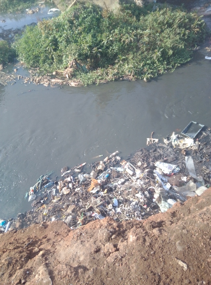The image depicts a segment of the heavily polluted Nairobi River in Nairobi, Kenya. The river's water is dark grey and turbid, showing surface ripples and scattered floating debris. On the far bank, dense green vegetation lines the water's edge. The immediate foreground shows a steep, exposed bank of reddish-brown earth. At the base of this near bank, a substantial accumulation of solid waste forms a distinct pile, extending into the water. This refuse consists primarily of discarded plastic bottles (clear, white, and blue), plastic bags, fabric remnants, and various household detritus. A dark, rectangular plastic object, possibly a crate, is partially submerged or resting within the trash accumulation on the near bank. No human figures are present. The scene illustrates significant environmental pollution with a large volume of waste accumulated along and within the waterway.