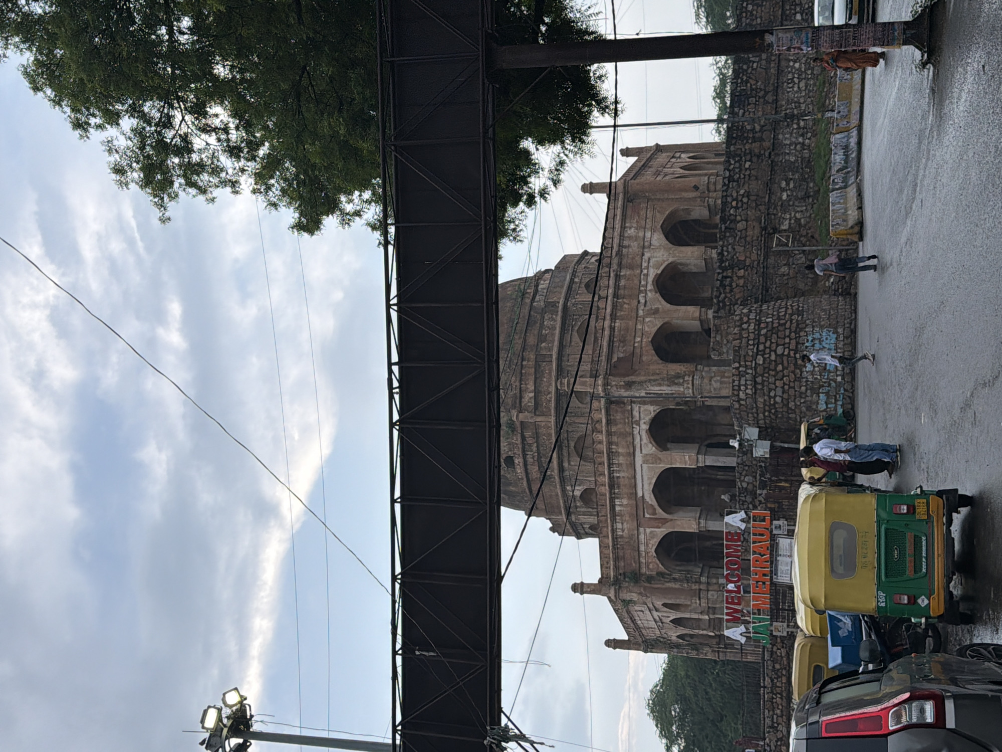 This image depicts a street scene in Delhi, India, likely during the day given the natural light. The weather appears overcast, possibly after rain, as the road surface is wet and reflective.
The central subject is a historical, domed structure with arched openings, constructed from rough stone. In front of this structure, a metal walkway or bridge spans across the street, partially obscuring the view of the building.
Several people are visible. Two individuals, one in a white shirt and the other in blue jeans, stand on the wet road near the historical building. Further down the street, another person in a white shirt is seen walking. A person in a colorful traditional Indian outfit stands near a pole, possibly selling something or waiting.
Three auto-rickshaws are parked on the side of the road. The foremost auto-rickshaw is yellow and green, with visible text on its rear. Another yellow auto-rickshaw is partially visible behind it, and a blue vehicle is behind that. A black car is parked in the foreground, its rear taillight clearly visible.
A prominent piece of text is displayed on a banner: "WELCOME JAI MEHRAULI". This suggests the location is Mehrauli, a historical area in Delhi known for its ancient ruins and monuments.
The overall scene conveys a sense of everyday life in a bustling Indian city, with a mix of historical architecture, modern transportation, and pedestrian activity. The wet road and overcast sky contribute to a muted, perhaps slightly somber, atmosphere.