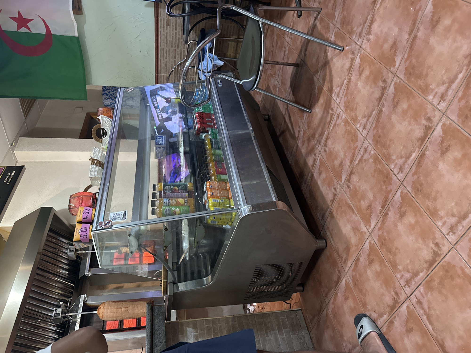 The image depicts the interior of a food establishment, likely a kebab shop or similar fast-food restaurant, possibly located in Alicante, Spain, given the presence of an Algerian flag. The scene is dominated by a refrigerated display case filled with beverages and what appears to be prepared food. Behind the display case, a vertical spit of meat, characteristic of a döner kebab, is visible, illuminated by red heating elements. The floor is tiled with terracotta-colored square tiles, showing signs of wear.
In the foreground, a person's left foot is visible, wearing a black and silver slide sandal. To the right of the display case, a metal chair with a black seat is positioned, and a small round table with a dark surface is partially visible in the background, with another metal chair tucked underneath.
The walls are a light color with a tiled section above the counter. The ceiling has a grid structure. Two price tags are affixed to the front of the display case, reading "5.00" and "4.30". The overall ambiance suggests a functional, no-frills eatery. The Algerian flag, green with a white crescent moon and star, hangs on the wall in the upper right quadrant of the image.