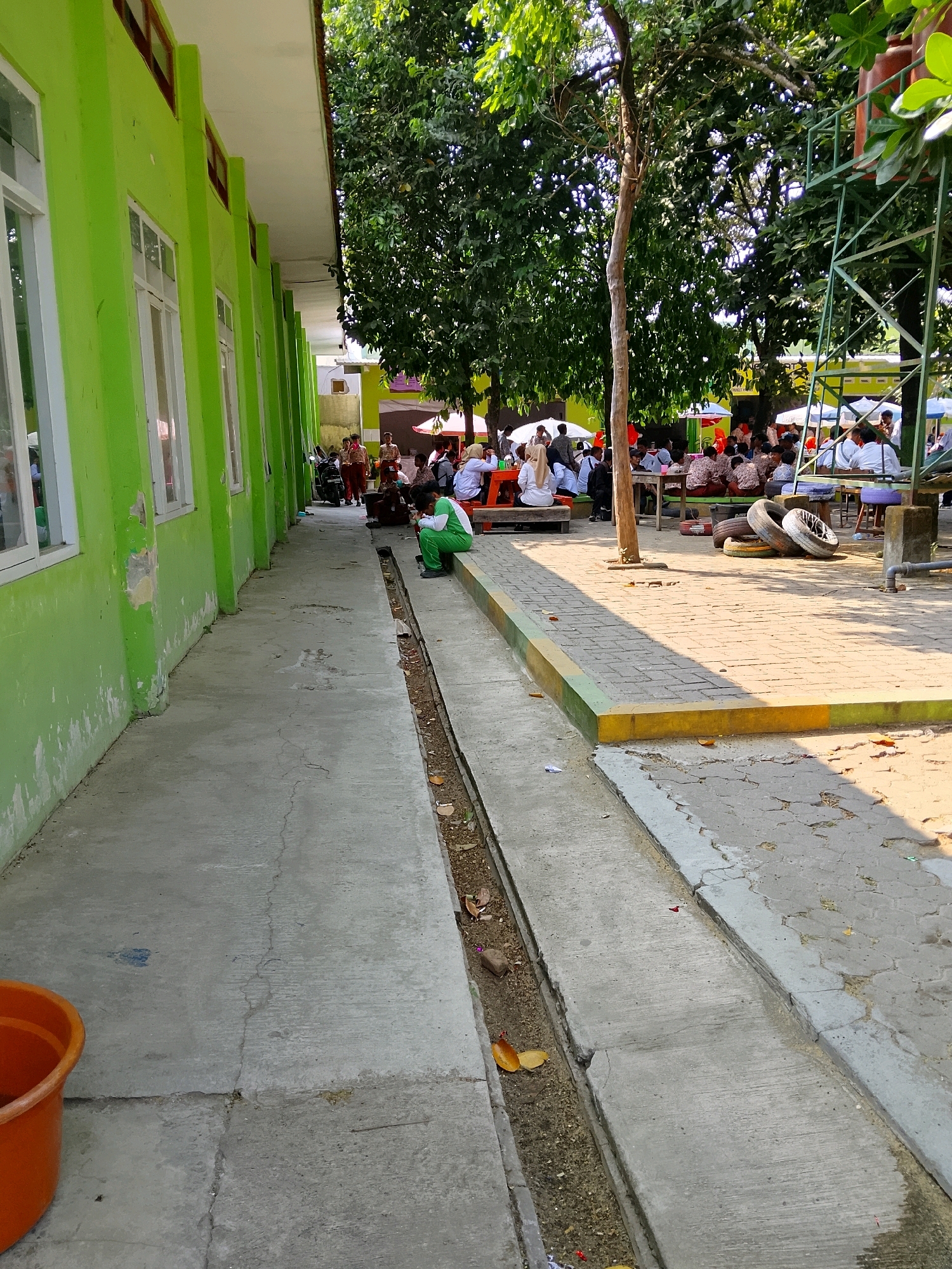 The image depicts an outdoor scene at a school in Kramat, Indonesia. On the left, a long, light green building with white-framed windows stretches into the distance. The building's wall shows some wear and tear near the bottom. A concrete walkway runs along the building, with a narrow, open drain on its right side. Further to the right, a paved area with interlocking bricks is visible, shaded by a large tree. In the background, a group of students are gathered at tables, some sitting and others standing, under umbrellas and trees. They appear to be wearing school uniforms, a mix of white and dark colors. To the far right, a green metal structure, possibly part of a playground, is partially visible. The lighting suggests it is daytime with the sun casting shadows.
