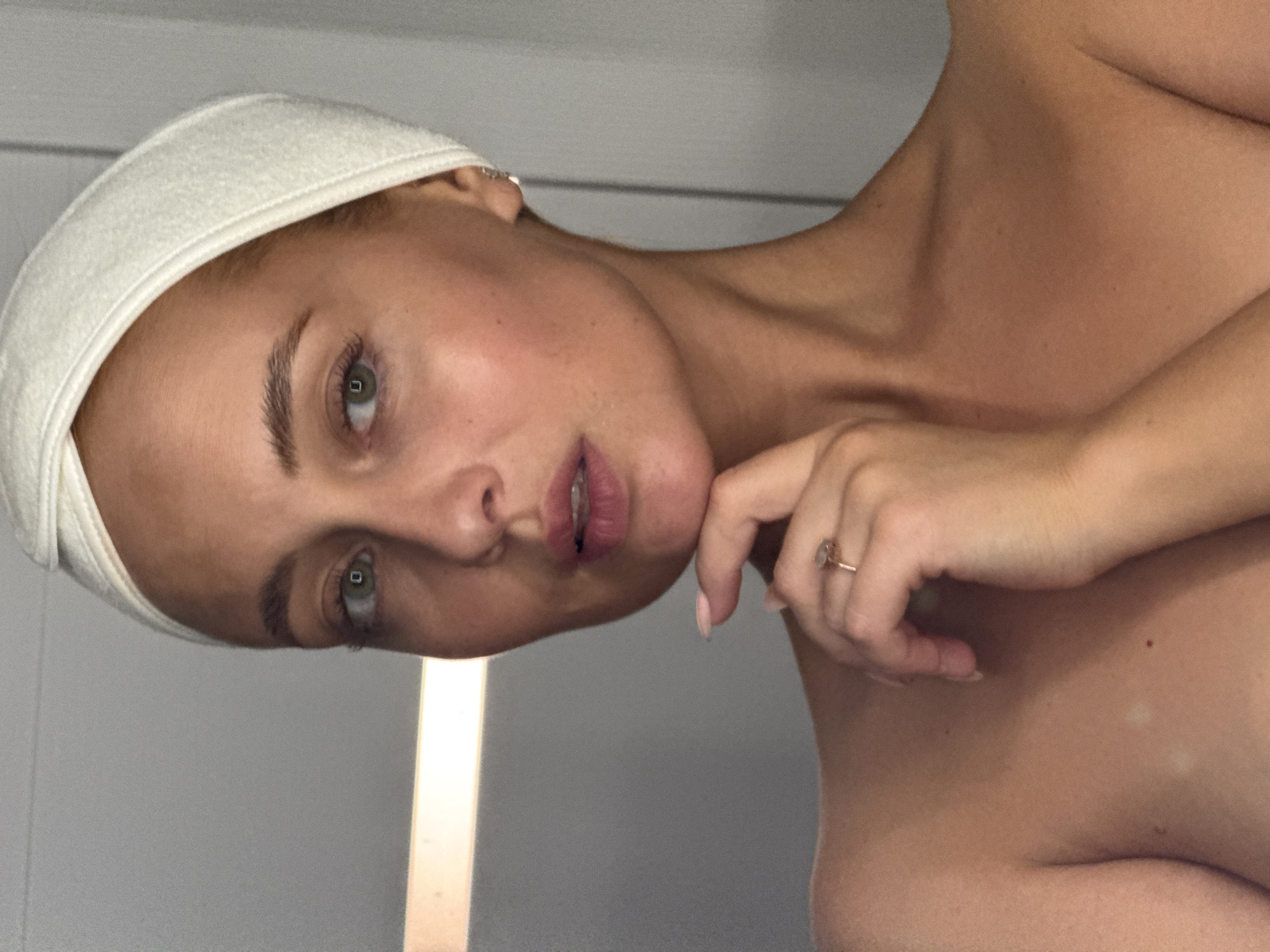 A woman with fair skin and light brown hair pulled back under a white terry cloth headband is posing for a close-up shot. Her head is tilted to the left, and her gaze is directed at the viewer. Her left hand is raised, with the index finger touching her chin. She is wearing a ring on her ring finger. The background appears to be a light-colored wall, possibly in an indoor setting. There is no indication of the location being Chaikovsky, Russia.