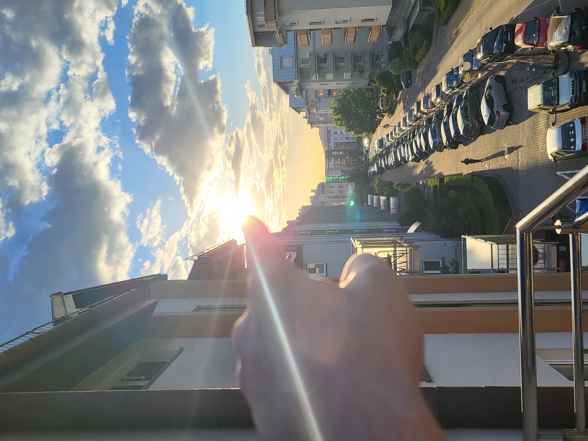 The image shows a view from a high angle, possibly a balcony, overlooking an urban scene. In the foreground, a hand is partially obscuring the view, with the index finger pointed towards the bright sun. The background consists of buildings with multiple stories and windows. The sky is visible, with a mixture of blue and clouds. A row of parked cars lines the side of the street near the buildings. Overall, the scene captures a moment during a sunny day in an urban environment.