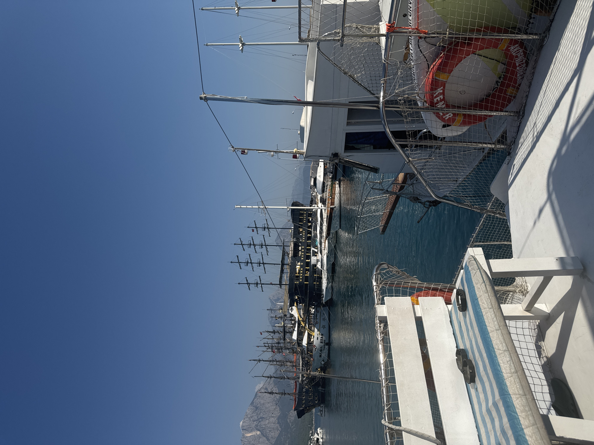 The image shows a harbor in Kemer, Turkey, filled with various boats and yachts. In the foreground, a portion of a white boat is visible, with a blue and white striped bench on its deck, equipped with several ashtrays. A red life preserver ring is also attached to the side of this boat. In the middle ground, several larger boats, including what appear to be pirate-themed ships with multiple masts, are docked. The water is calm and reflects the blue sky. In the background, a range of rugged mountains with some sparse vegetation can be seen, with a clear blue sky overhead. The scene depicts a sunny day in a tourist-friendly coastal town.