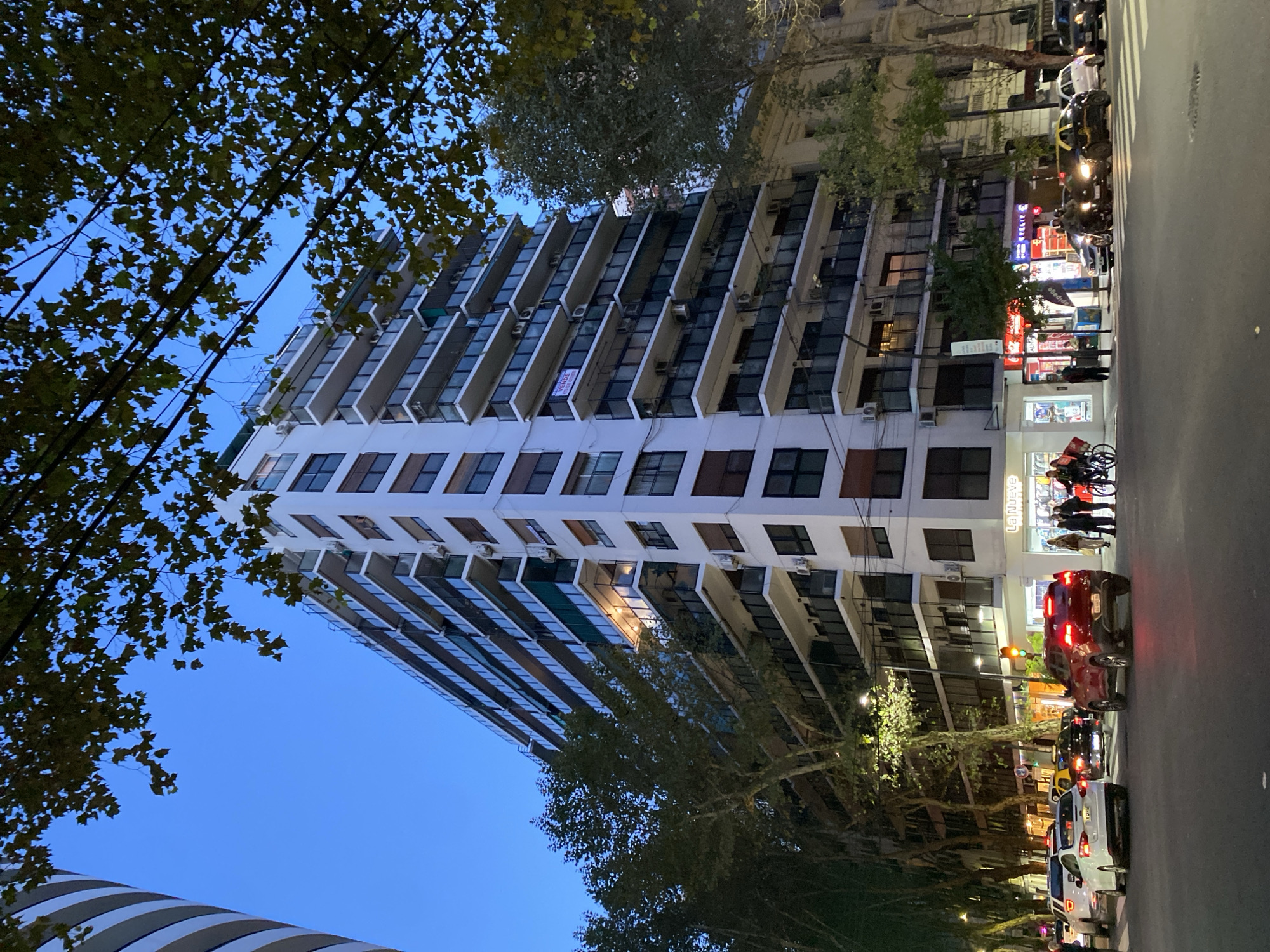 The image shows a tall, white apartment building in Buenos Aires, Argentina, viewed from street level. The building has many balconies and windows. Trees are visible on either side of the building, partially obscuring the view. Street lights illuminate the street, which is lined with cars and pedestrians. The time of day appears to be dusk or evening. A business with a red sign is next to the building. People are gathered near the store. There are also people on bicycles.
