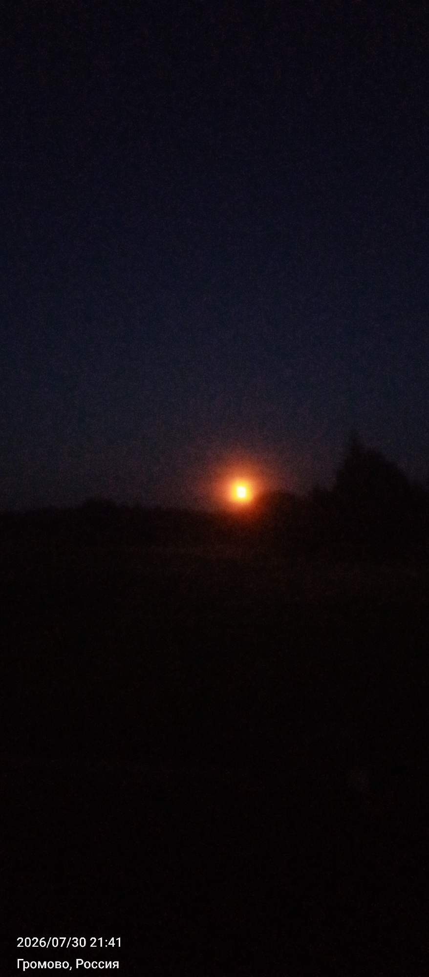 The image depicts a dark sky with a bright, blurred orange sun near the horizon. The foreground is extremely dark, obscuring any details of the landscape. Silhouetted against the faint light of the sun are dark shapes that appear to be trees or foliage. The bottom left corner of the image contains text indicating the date and time as "2026/07/30 21:41" and the location as "Громово, Россия" (Gromovo, Russia).