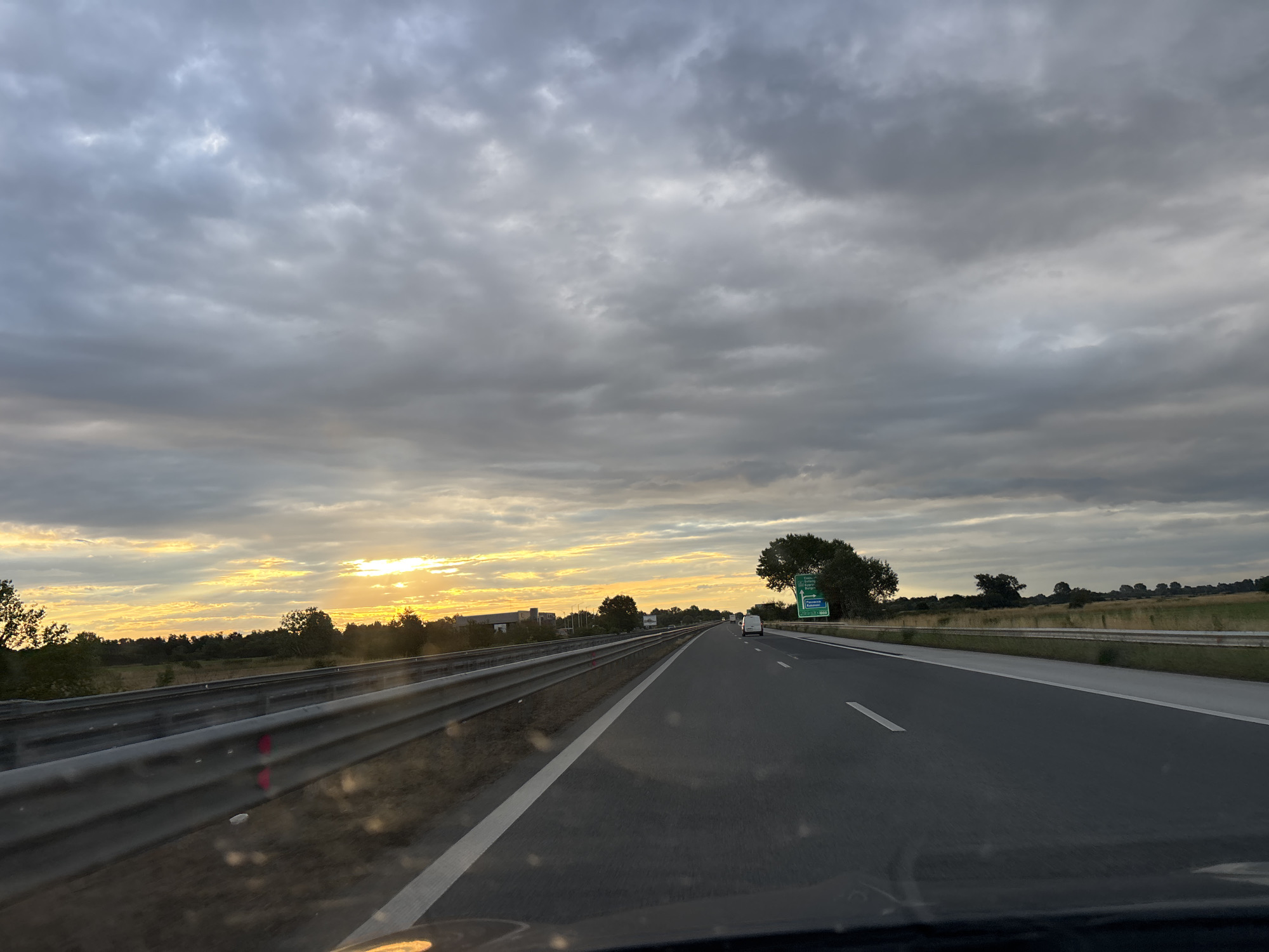 This image depicts a highway scene at sunset, taken from the perspective of a vehicle driving on the road. The sky is filled with dramatic clouds, with a sliver of bright orange and yellow light from the setting sun visible on the horizon. The highway is paved and appears to be multi-lane, with a metal guardrail running along the left side. The right side of the road features a grassy verge and a few scattered trees. In the distance, there are more trees and some low-lying buildings. A green highway sign with white lettering is visible on the right side of the road, indicating directions to various locations including "Skutare". There are a few vehicles on the road in the distance, and no people are directly visible. The overall impression is one of travel on a highway under a cloudy, colorful sky.