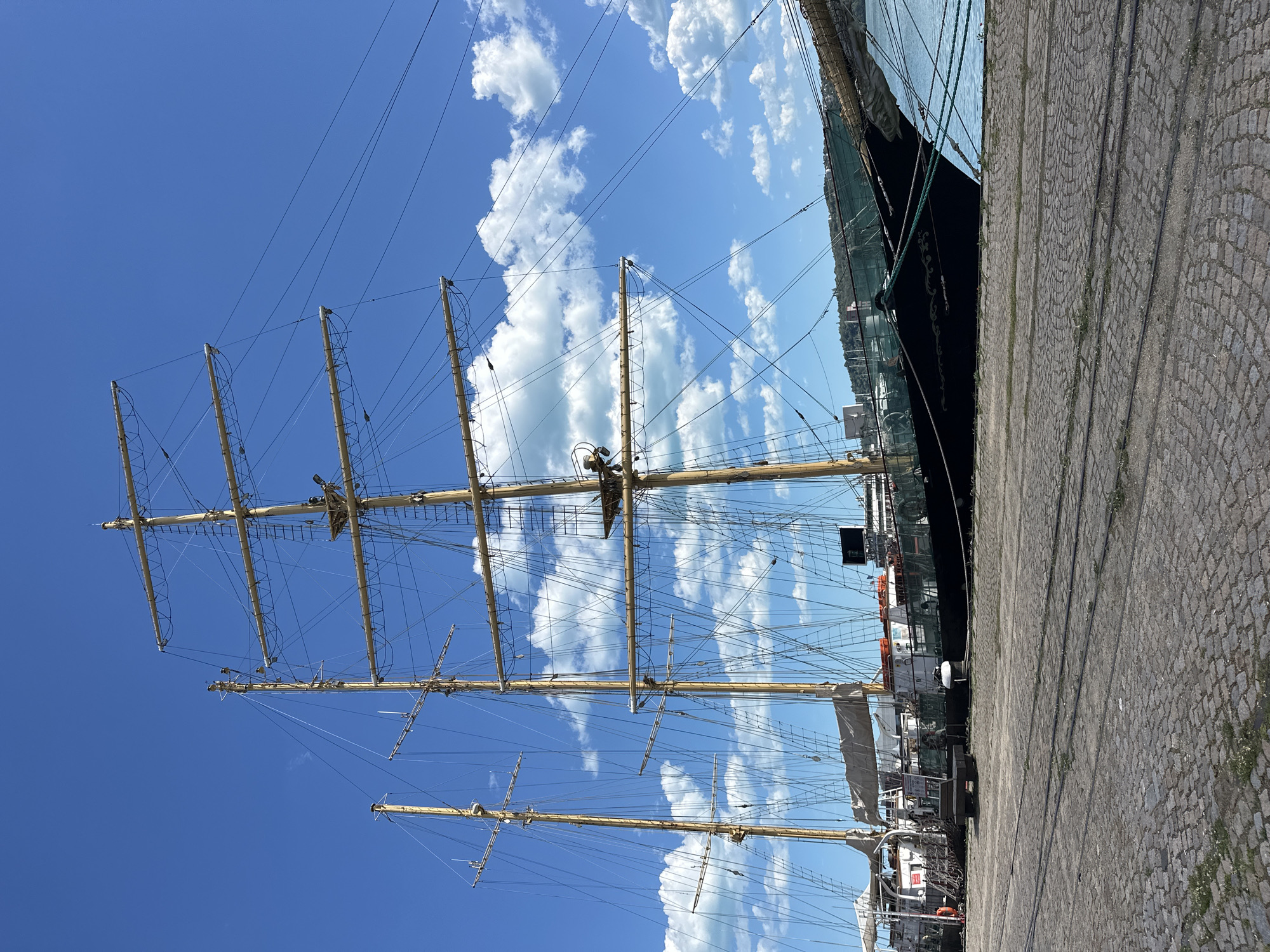 The image depicts a tall sailing ship docked in Varna, Bulgaria, with its masts and rigging prominently displayed against a backdrop of blue sky with scattered clouds. The ship's dark hull is alongside a stone-paved quay. The masts are tall and thin, reaching high into the sky, with complex rigging and spars. Other boats are docked near this tall ship in the background. The sky overhead is bright blue with a mix of puffy cumulus clouds.