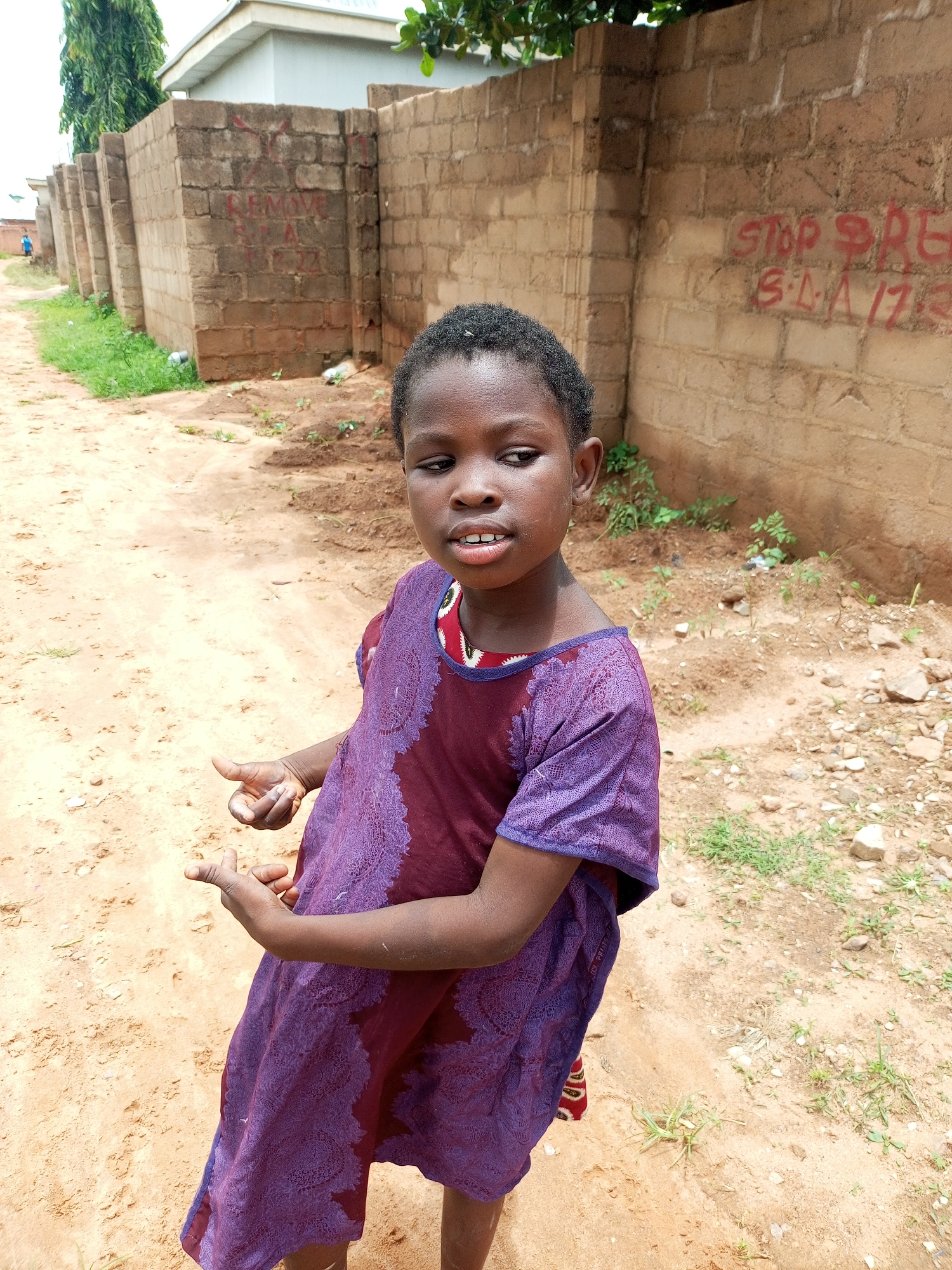 In Tempure, Nigeria, a young girl stands in a dirt lot next to a brick wall covered in graffiti. She wears a purple dress adorned with a floral pattern. Her hands are raised, and she appears to be speaking or gesturing. Behind her, the brick wall is marked with red spray paint, some of which is legible, reading "STOP PRE" and possibly "S.L.A. 17:3" or similar. A small section of a building and some trees are visible in the background, along with sparse green vegetation growing around the wall. The ground is predominantly sandy dirt with scattered rocks and debris.