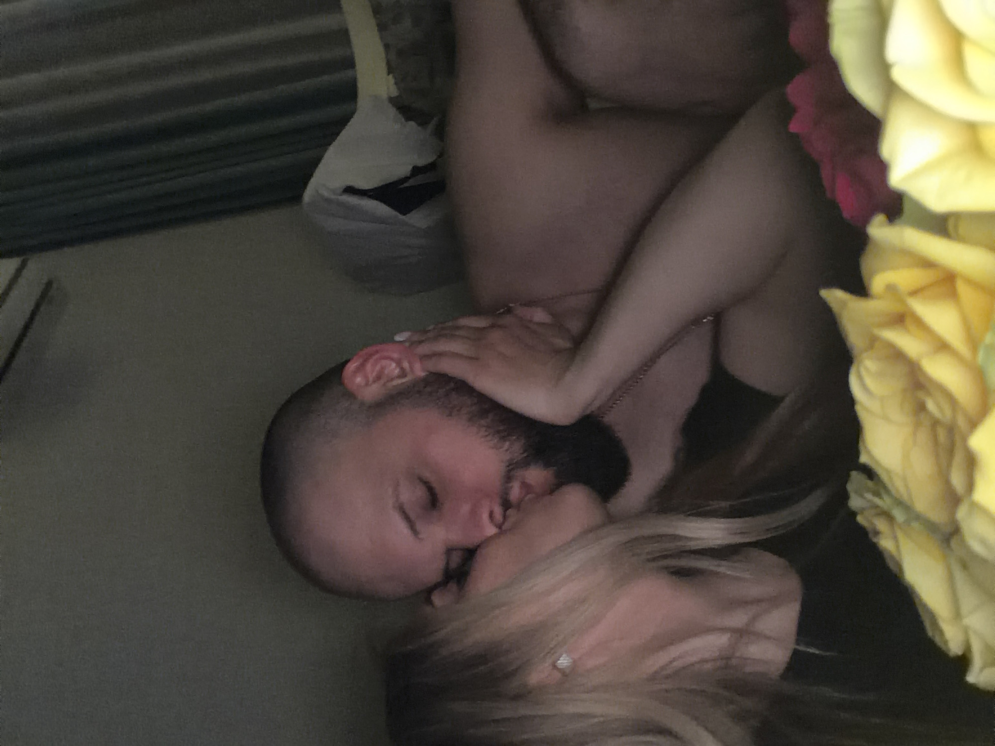 A man and a woman are kissing. The man has a shaved head and a beard. The woman has blonde hair and is wearing a ring. There are yellow and red roses in the foreground. A plastic bag is in the background. The location is not discernible as being in Votkinsk, Russia.