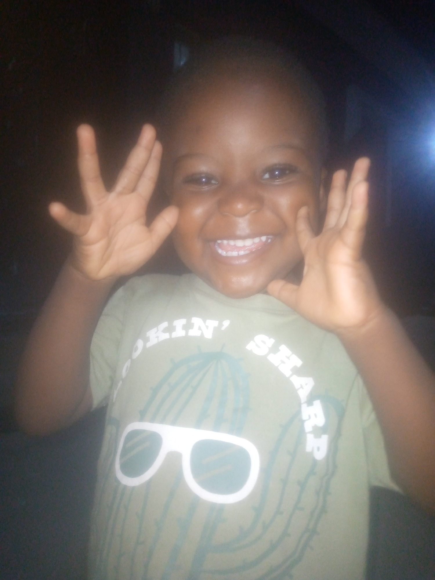 A child with dark skin is smiling and holding up both hands with fingers spread, facing the camera. The child is wearing a light green T-shirt with the words "LOOKIN' SHARP" printed above a stylized cactus and sunglasses design. The background is dark and indistinct. The location is Unknown, Unknown.
