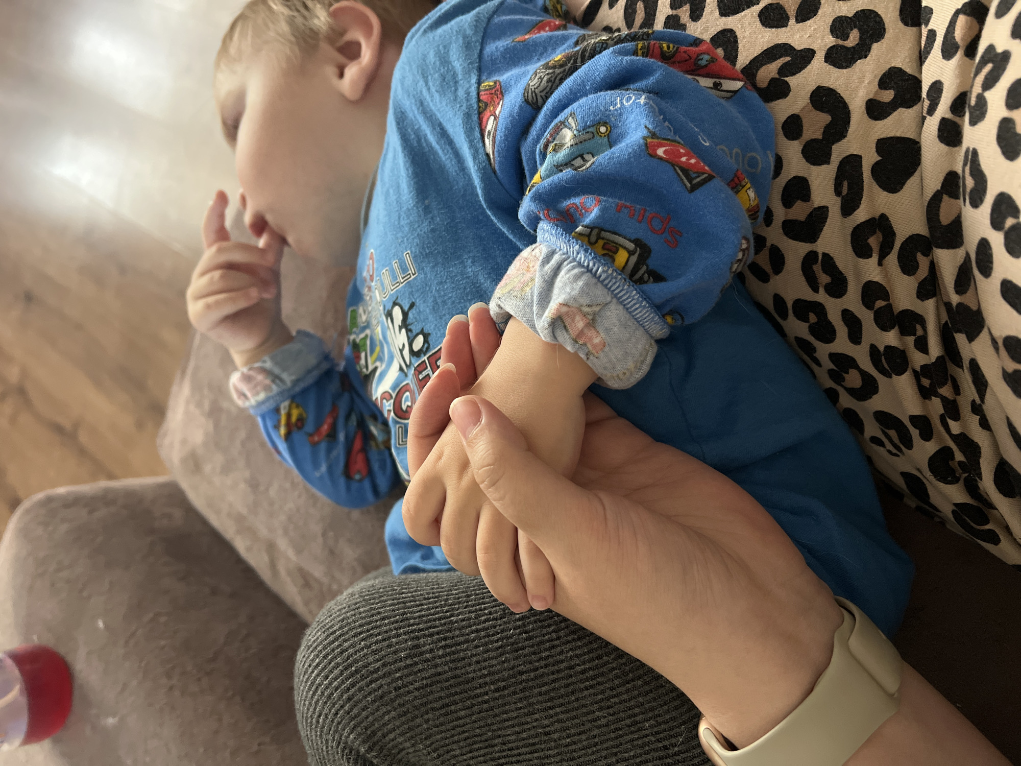 In an indoor setting, a person's hands are clasped around a child's hand. The child, who appears to be a toddler, is wearing a blue long-sleeved shirt with a vehicle pattern. The child is lying down, with their head resting on a surface that is partially visible. The surrounding environment includes a soft, textured surface, likely a sofa or bed, and a wooden floor. A partially visible red-topped container is also present. The scene takes place in Noviy, Russia.