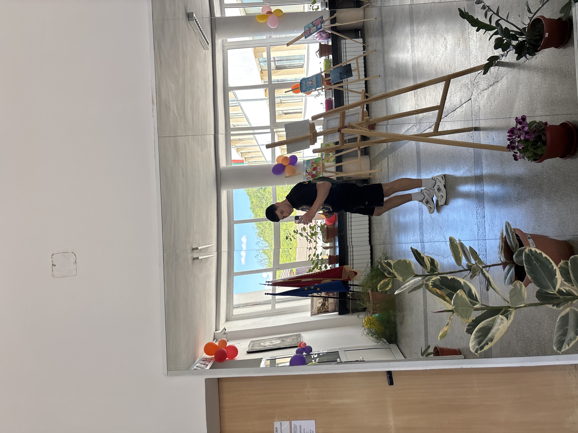A young man in black shorts and a black t-shirt stands in an indoor exhibition space. He is looking at a phone in his hands. Wooden easels display artwork. Balloons are attached to the window frames. There are potted plants in the foreground and background. Flags are near the windows. The scene is in Zlatograd, Bulgaria.
