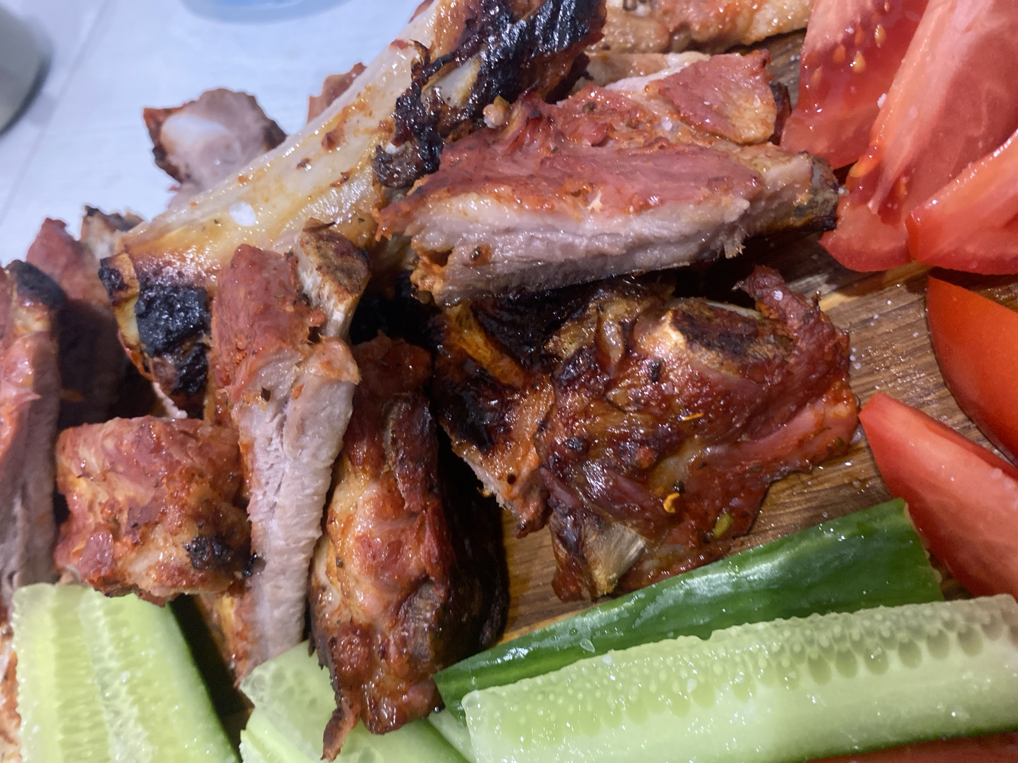 The image depicts a close-up shot of food presented on a wooden board. The main focus is on grilled ribs, with a portion of them displaying bones. Complementing the ribs are sliced cucumbers and tomatoes, arranged around the meat. The ribs are charred in some areas, indicating a cooking method involving high heat. The cucumbers appear fresh and are cut into elongated pieces. The tomatoes are ripe and have been sliced into wedges. The presentation of the food suggests a meal setting, possibly outdoors. No people are visible in the frame. There is no information about the location in the city of Chaikovsky, Russia.