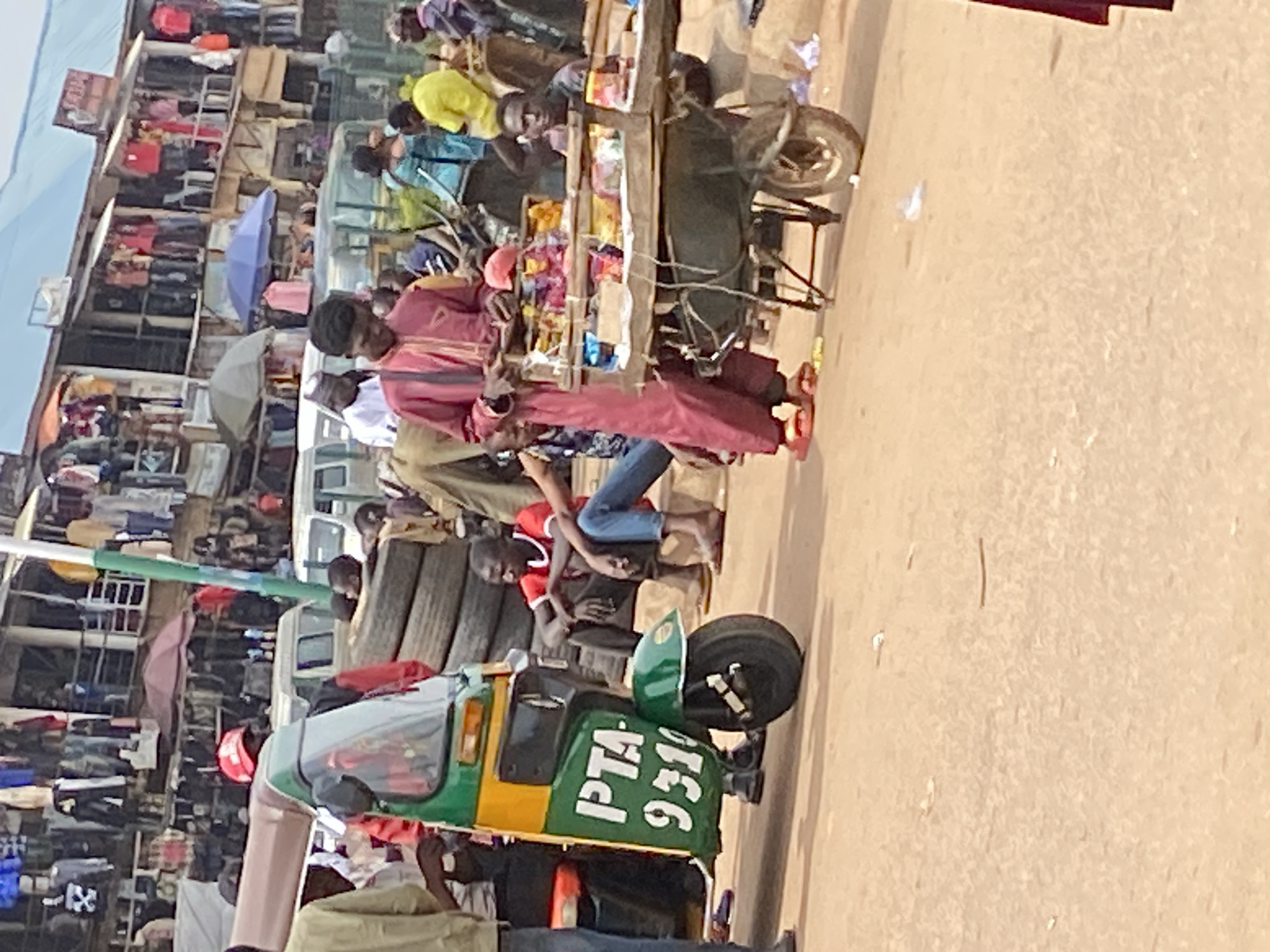 In Bukuru, Nigeria, a bustling street scene unfolds. A green and yellow motorized tricycle, identified with the markings "PTA - 9319", is parked on a dusty, unpaved road. A man in a red jacket and red cap stands beside the tricycle, looking towards the left. Behind him, a stack of dark tires is visible. To the right of the tricycle, a man in a maroon outfit stands near a wooden cart filled with various items, possibly snacks or small goods. He is looking down at the cart and appears to be interacting with it. Several other people are present in the background, some sitting and others standing. The environment suggests a market or roadside commercial area, with buildings lining the street and merchandise displayed. The lighting indicates it is daytime.