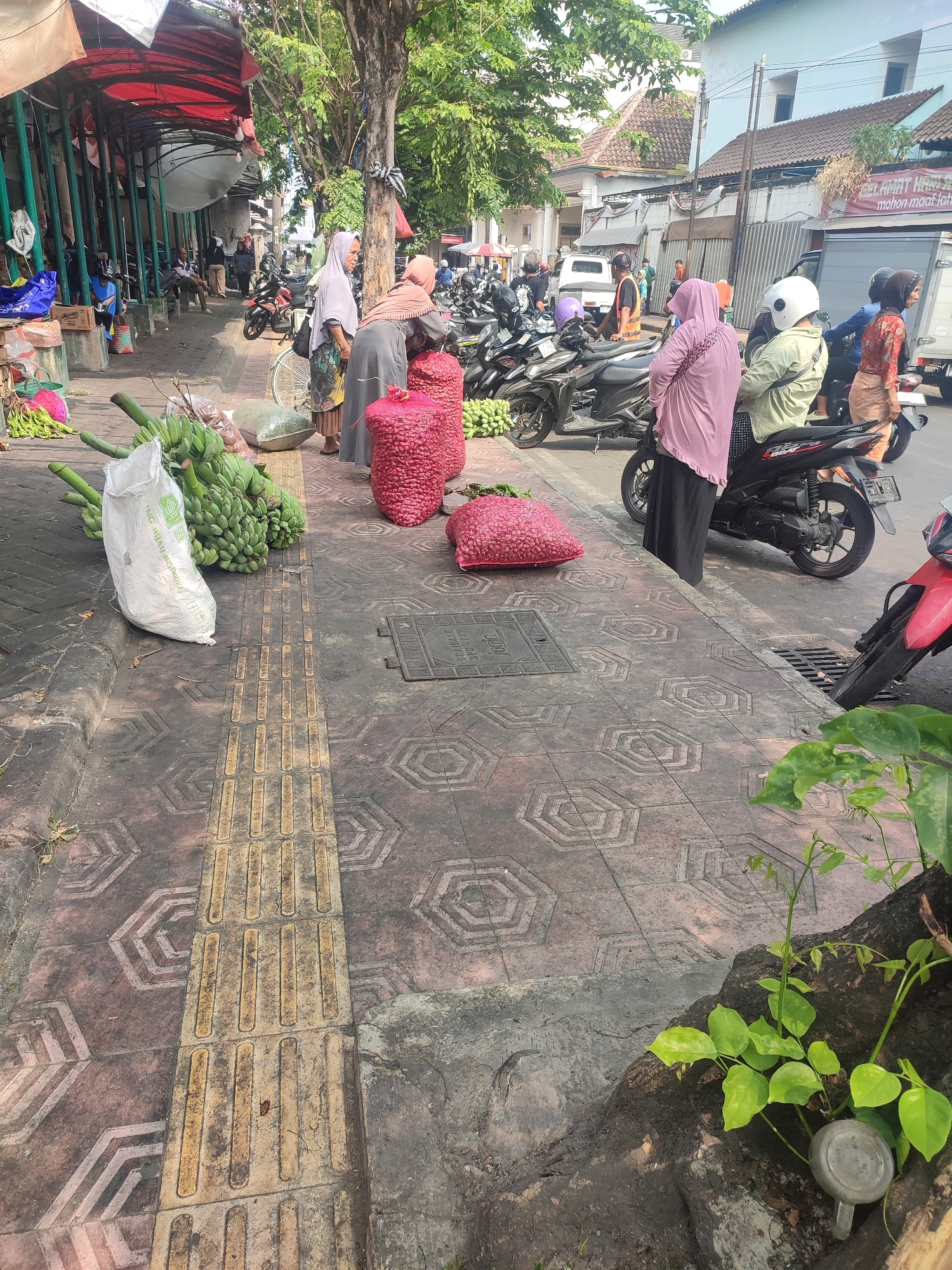 The image depicts a street market scene in Yogyakarta, Indonesia, characterized by a sidewalk with tactile paving for the visually impaired. On the sidewalk, several people are present, most of whom are wearing hijabs. In the foreground, large sacks filled with red produce, possibly onions or chilies, are displayed. To the left, bunches of bananas are arranged, some in a white sack. Beyond the produce, stalls with red awnings are visible, with more people and parked motorcycles. In the background, a white building and trees line the street. The overall impression is of a busy, outdoor market environment.