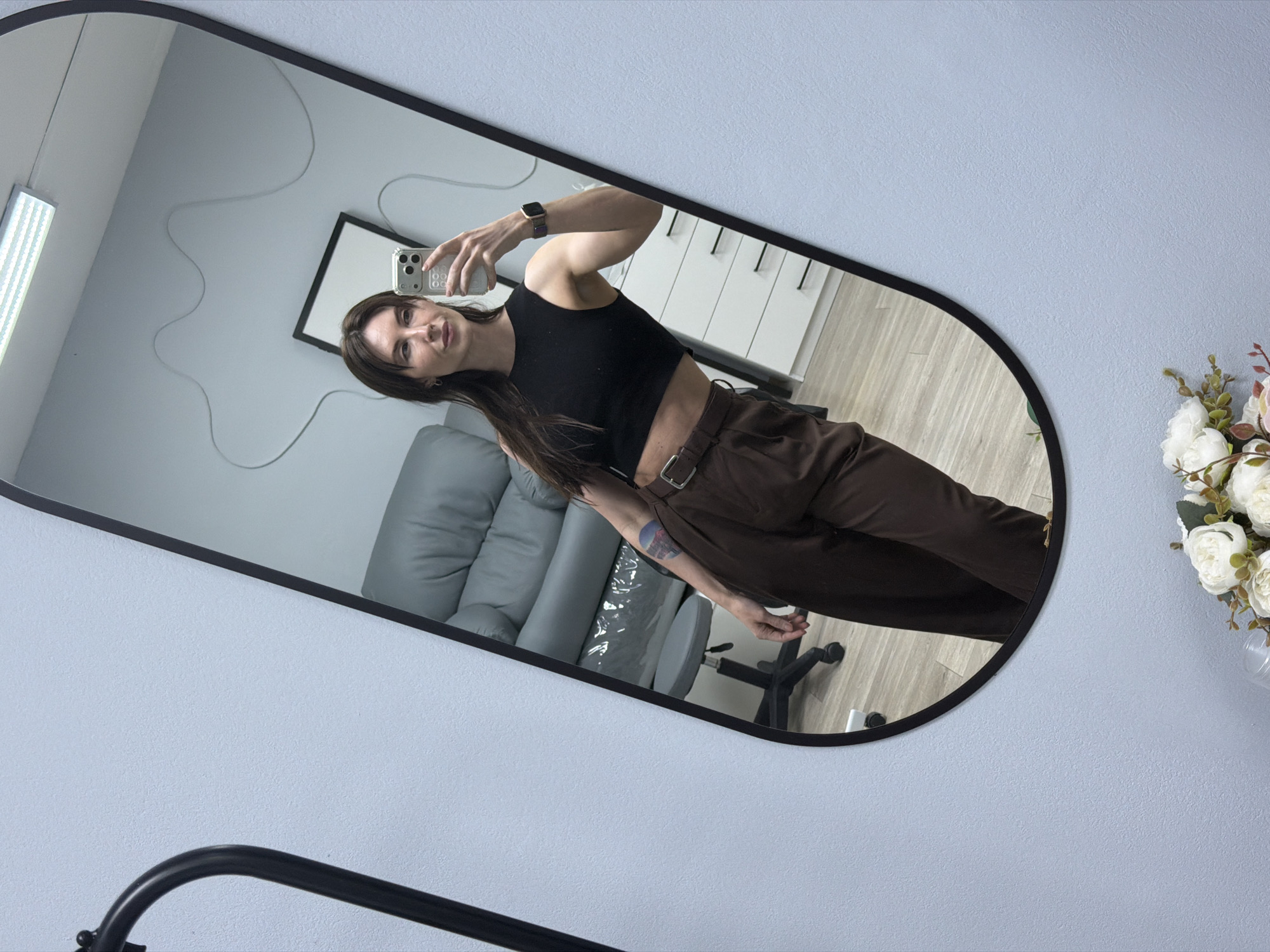 A woman is taking a selfie in a mirror. The mirror is oval-shaped with a black frame, hanging on a light blue wall. She is wearing a black crop top and brown pants with a belt. A watch is visible on her left wrist. The reflection in the mirror shows a gray couch, a desk chair, and a white dresser. A black-framed picture hangs on the wall in the background. A lamp is visible on the ceiling. A vase of white flowers is situated in the bottom right corner of the image. The location may be in Perm, Russia.