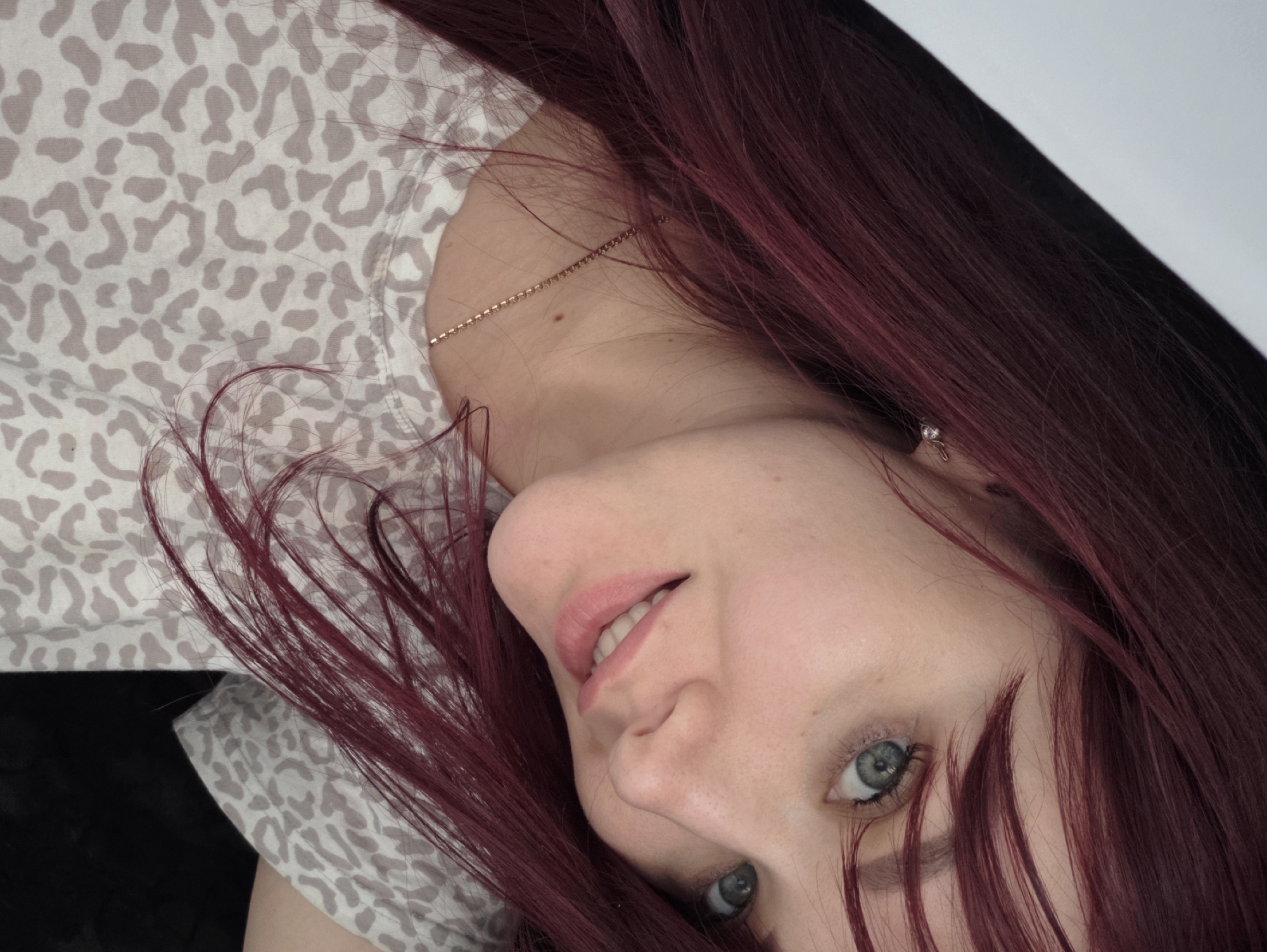 The image is a close-up, selfie-style photograph of a young woman with long, dark red hair. She has light blue eyes and fair skin with freckles. She is wearing a t-shirt with a light gray and white leopard print pattern and a delicate gold chain necklace. A small, sparkly earring is visible in her right ear. She is smiling softly, and her head is tilted to the side, revealing her face and upper torso. The background is dark, with a sliver of bright white on the upper left, suggesting the presence of a light source or reflective surface. There are no specific landmarks or environmental details visible that would indicate a location within the city of Chaikovsky, Russia, or any other specific city.