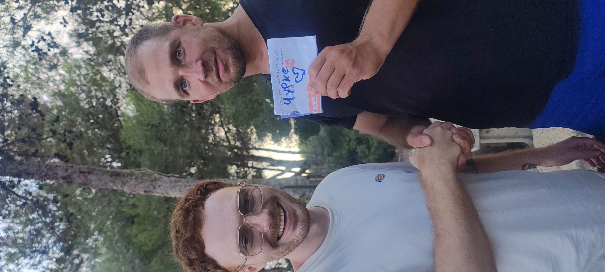 The image depicts two men standing outdoors, likely in Nesher, Israel, given the context. The man on the left has red hair, a beard, and is wearing glasses and a white t-shirt with a Dickies logo. He is smiling and shaking hands with the man on the right. The man on the right has short brown hair and is wearing a black t-shirt. He is holding an envelope with writing and a heart drawn on it in blue. The background consists of trees and foliage, suggesting a natural or park-like setting. The lighting indicates it is daytime.