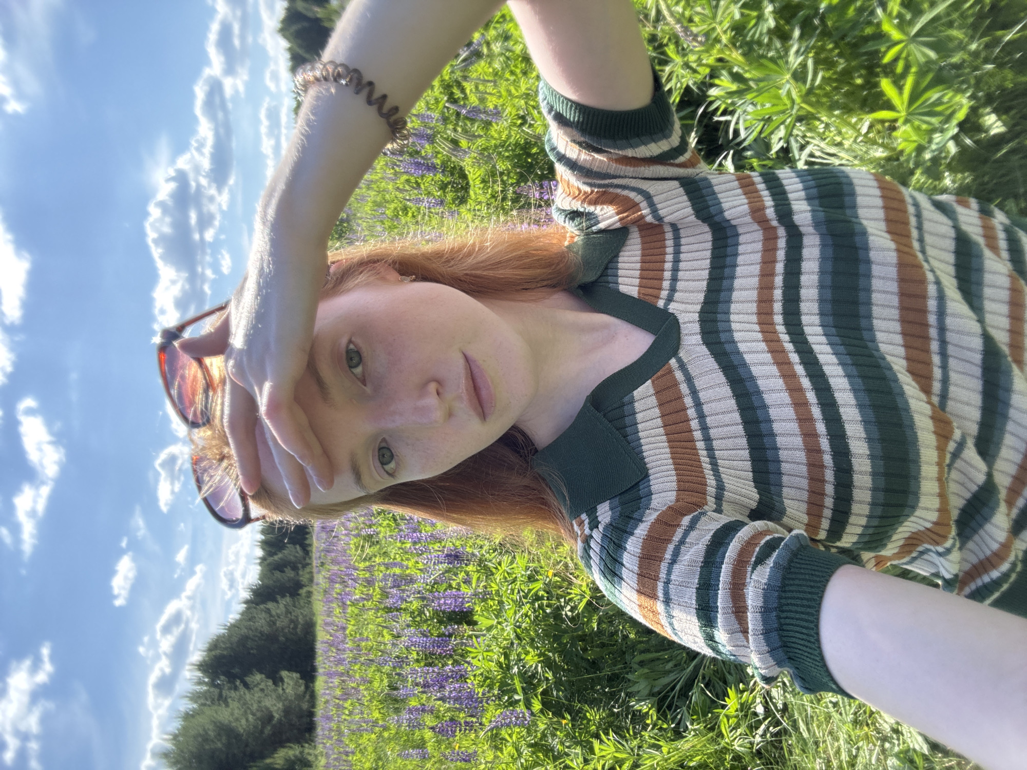 In Chaikovsky, Russia, a young adult female with red hair and visible freckles poses for a selfie outdoors. She wears a ribbed knit polo shirt featuring horizontal stripes in dark green, brown, light blue, and off-white. Sunglasses with reddish-brown frames are pushed onto the top of her head. Her right hand is raised to her forehead, shielding her eyes from direct sunlight. A coiled, translucent brown hair tie or bracelet is on her left wrist. The subject is situated within a vibrant field of tall green foliage interspersed with numerous purple lupine flowers. In the background, a dense line of coniferous trees forms a forest boundary beneath a clear blue sky dotted with scattered white cumulus clouds. The scene is brightly illuminated by natural daylight.