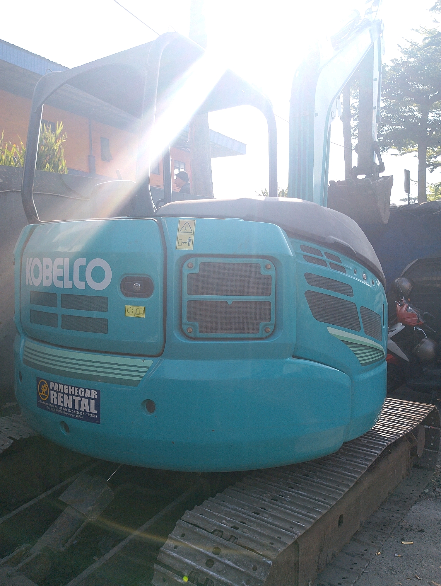 An excavator, a Kubota brand, painted in a teal color, is parked outdoors. The excavator has a Kobelco logo on its side, and a "Panghegar Rental" sticker below it. The excavator is sitting on a tracked base. In the background, a building with an orange facade and blue roof can be seen. There are trees in the distance. The sun is shining brightly, creating lens flare across the image. The scene appears to be a rental yard or construction site in Lembang, Indonesia.