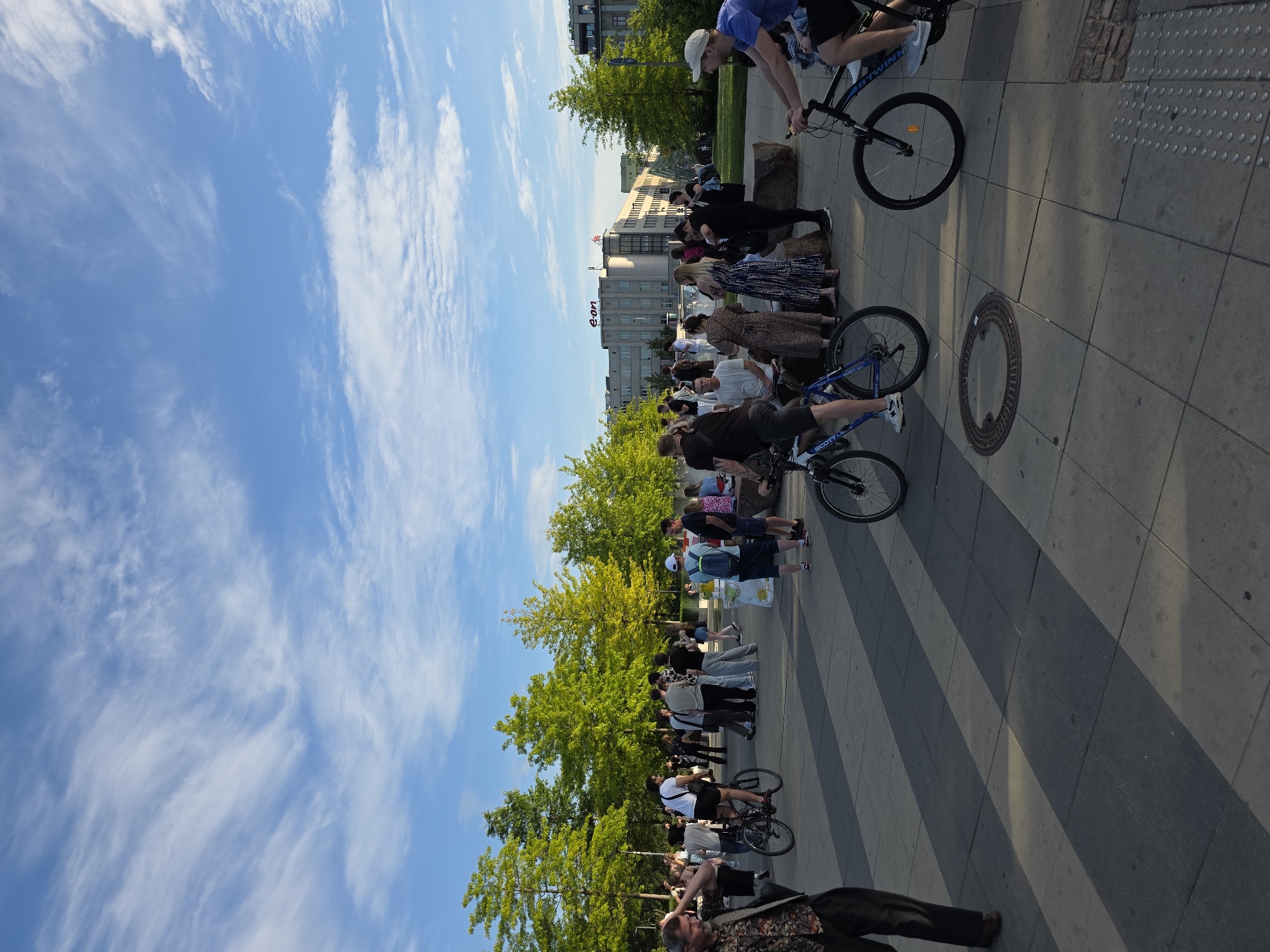 The image shows a group of people standing in a line on a paved walkway. They appear to be observing something or waiting for an event. Several individuals are on bicycles. The scene is outdoors with a bright blue sky filled with wispy clouds, suggesting a pleasant day, likely afternoon due to the long shadows cast by the trees and people. Lush green trees line the walkway, and in the background, there's a modern building with the text "e.on" prominently displayed. The atmosphere seems calm and orderly. The people are dressed in casual clothing, and their postures don't suggest any particular emotion, other than general attentiveness.