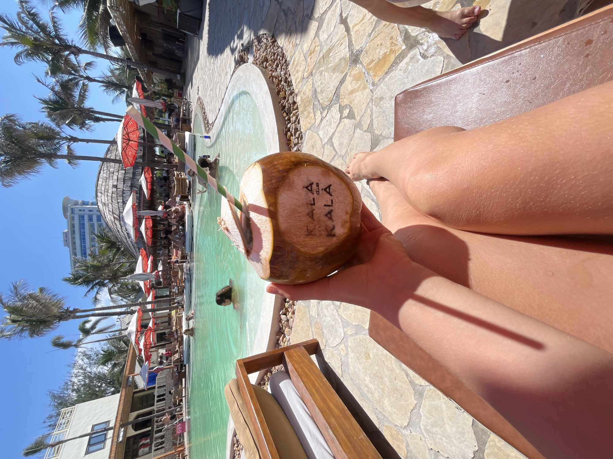 The image captures a vibrant, sun-drenched scene at a tropical resort or beach club, likely in Đà Nẵng, Vietnam. In the foreground, a person's tanned legs and hands are visible as they recline on a brown lounge chair. They hold a fresh young coconut with a green and white striped straw, displaying the stamped logo "KALA KALA CLUB" on its side.
The central feature of the scene is a large, irregularly shaped swimming pool with clear, light green water. Surrounding the pool are numerous lounge chairs shaded by red and white striped umbrellas, where other guests are relaxing. Several individuals are visible in the pool, swimming or wading. The ground around the pool is paved with light-colored, irregularly shaped stone tiles, featuring a decorative border of small pebbles.
Numerous tall palm trees are scattered throughout the resort area, enhancing the tropical ambiance. In the background, a multi-story modern building, likely a hotel, stands under a clear blue sky, contrasting with a more rustic, thatched-roof structure closer to the pool, possibly a bar or restaurant.
The bright sunlight, casting distinct shadows, indicates it is midday or early afternoon on a clear, warm day. The overall activity depicted is one of relaxation and enjoyment at a popular vacation destination, with people unwinding by the pool. The visible text on the coconut is "KALA KALA CLUB".