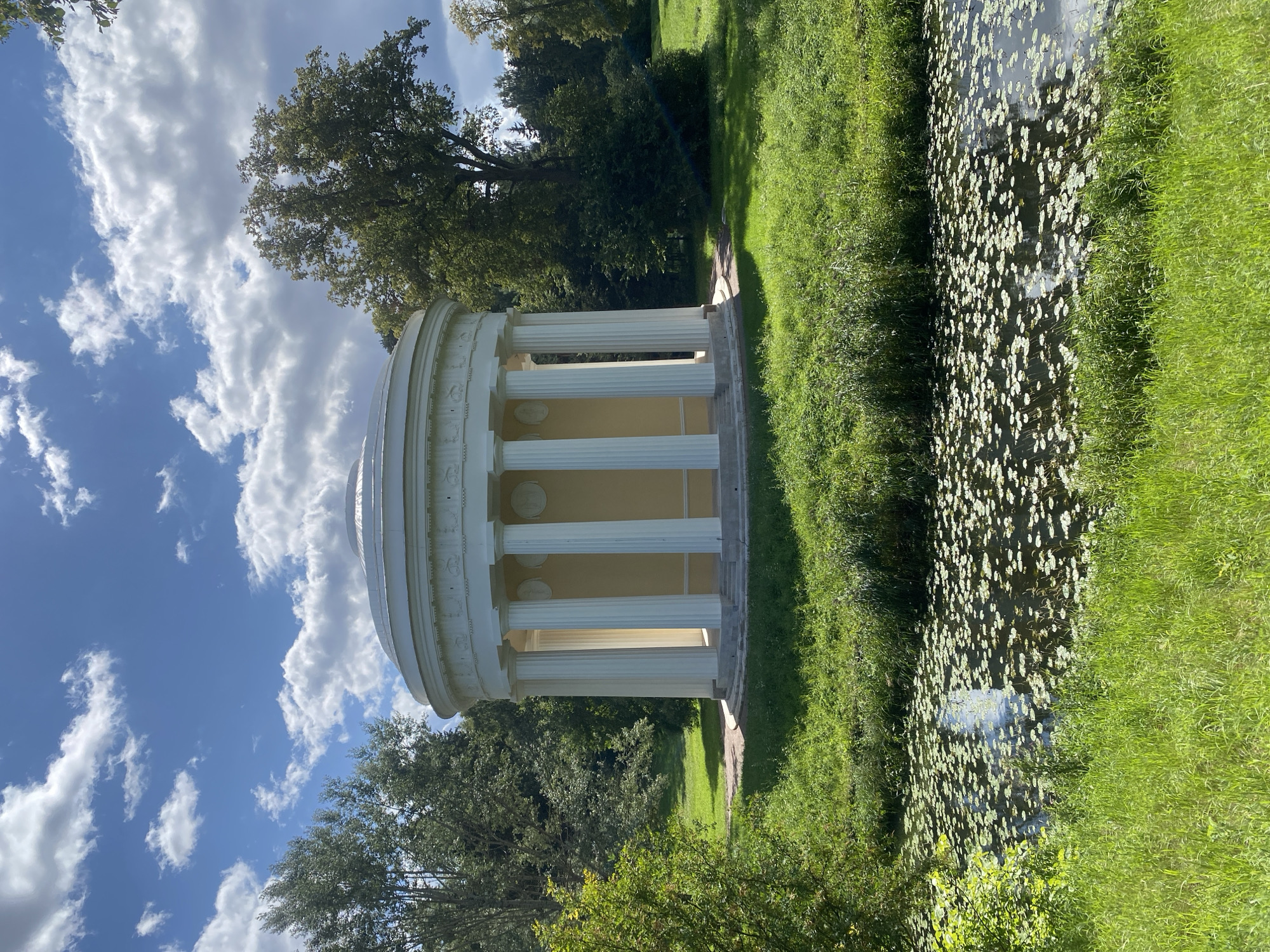 The image shows the Temple of Apollo at the Pavlovsk Park in Pavlovsk, Russia. The circular neoclassical building, painted a pale yellow with white columns, is situated in a lush green park with tall trees. In the foreground, a body of water is covered with lily pads. The sky is blue with scattered white clouds. The scene is bathed in sunlight, casting shadows on the grass. No people or animals are visible in the image.