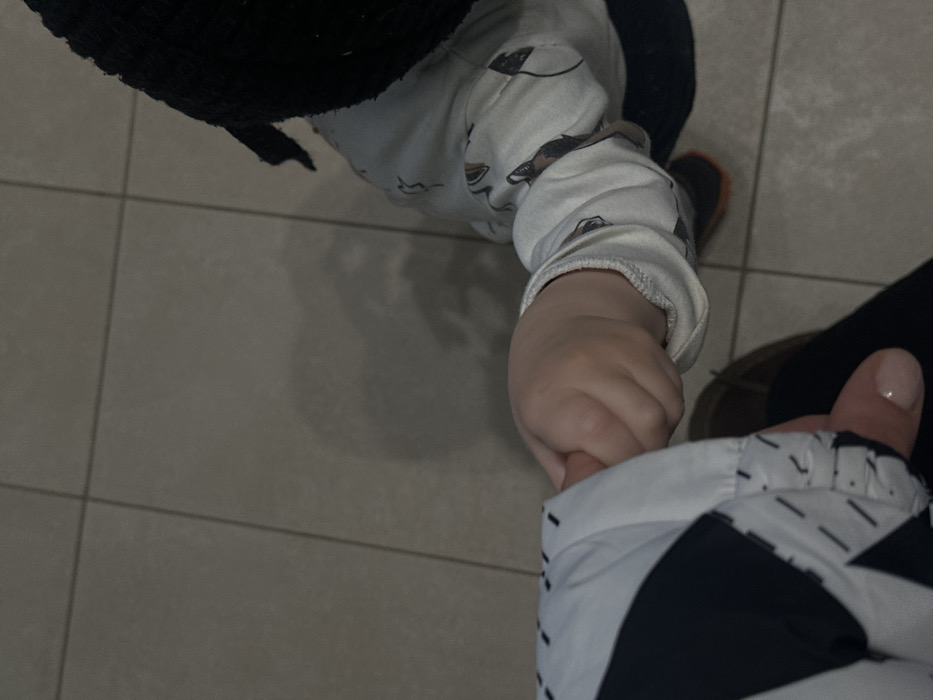 The scene, located in Ivashevo, Russia, depicts a top-down view of two individuals, an adult and a child, interacting in an indoor environment. The adult, positioned to the lower left, wears a jacket featuring a black and white geometric pattern with dashed line details near the cuff, and a dark garment, possibly pants. A part of a brown shoe is visible. The adult's hand, exhibiting light pink manicured fingernails, is visible in the lower left quadrant. The child, positioned to the lower right, wears a white long-sleeved top adorned with a repeating pattern of dark, stylized figures. A dark, ribbed knit hat covers the child's head, and an orange and dark-colored shoe is partially visible on the right. The primary action shows the child's right hand grasping the adult's left index finger or thumb. The environment consists of a light gray tiled floor with visible darker grout lines, suggesting an institutional or commercial setting. Overhead lighting casts soft shadows, particularly from the child onto the floor. No further identifying architectural or contextual details are discernible.