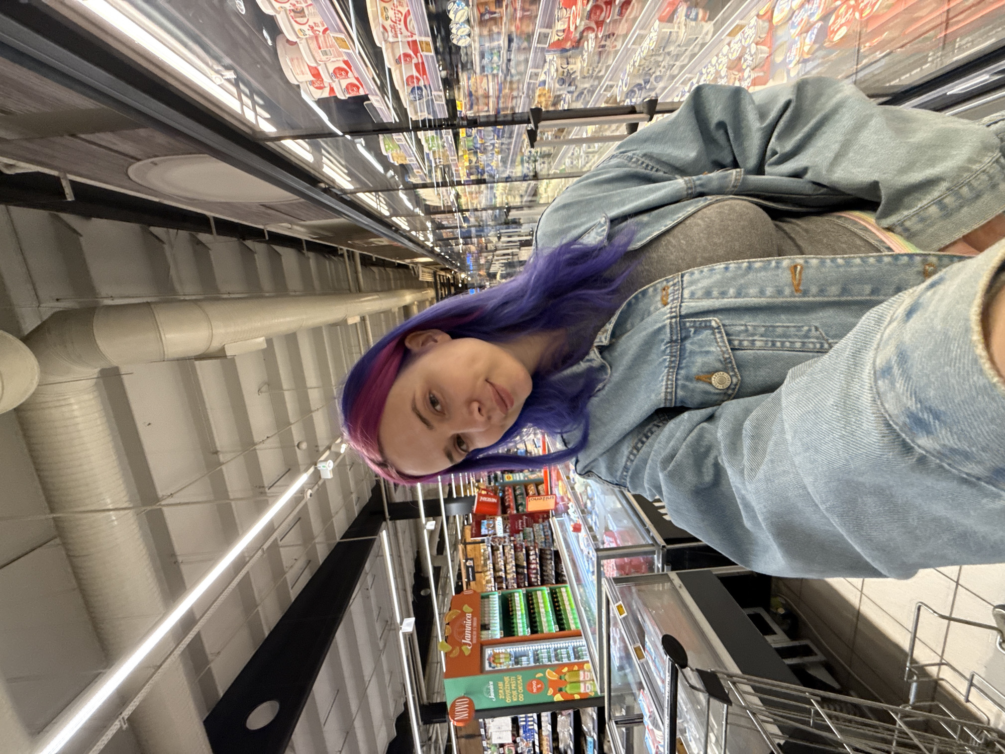 A woman with purple and pink hair wearing a denim jacket stands in the aisle of a grocery store. She is holding a shopping cart. The aisle is filled with shelves stocked with various food and beverage items. A sign for "Jamaica" brand juice is visible. The image was taken in City of Zagreb, Croatia.