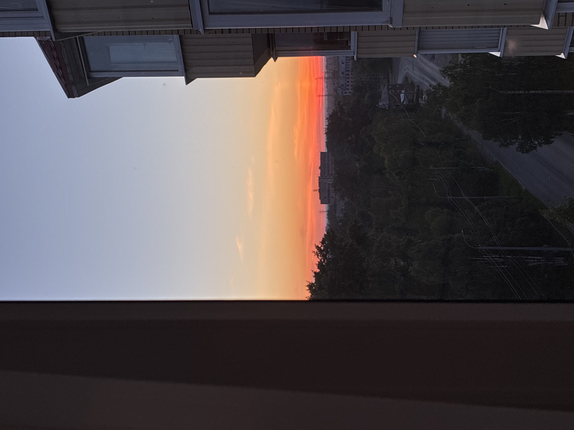 The image is a medium-shot, eye-level, vertical photograph taken from a window overlooking a city at sunset. The sky is a gradient of blue to orange, with a bright red band on the horizon. In the foreground, on the right side of the frame, is a portion of a light-colored building with vertical paneling and several windows. The left side of the frame is dominated by a dark, vertical structure, possibly the interior wall of the room from which the photo is taken. In the mid-ground, a dense canopy of dark green trees lines a winding road. Beyond the trees, several industrial-looking buildings and power lines are visible. The overall impression is one of a city transitioning from day to night under a dramatic sunset.