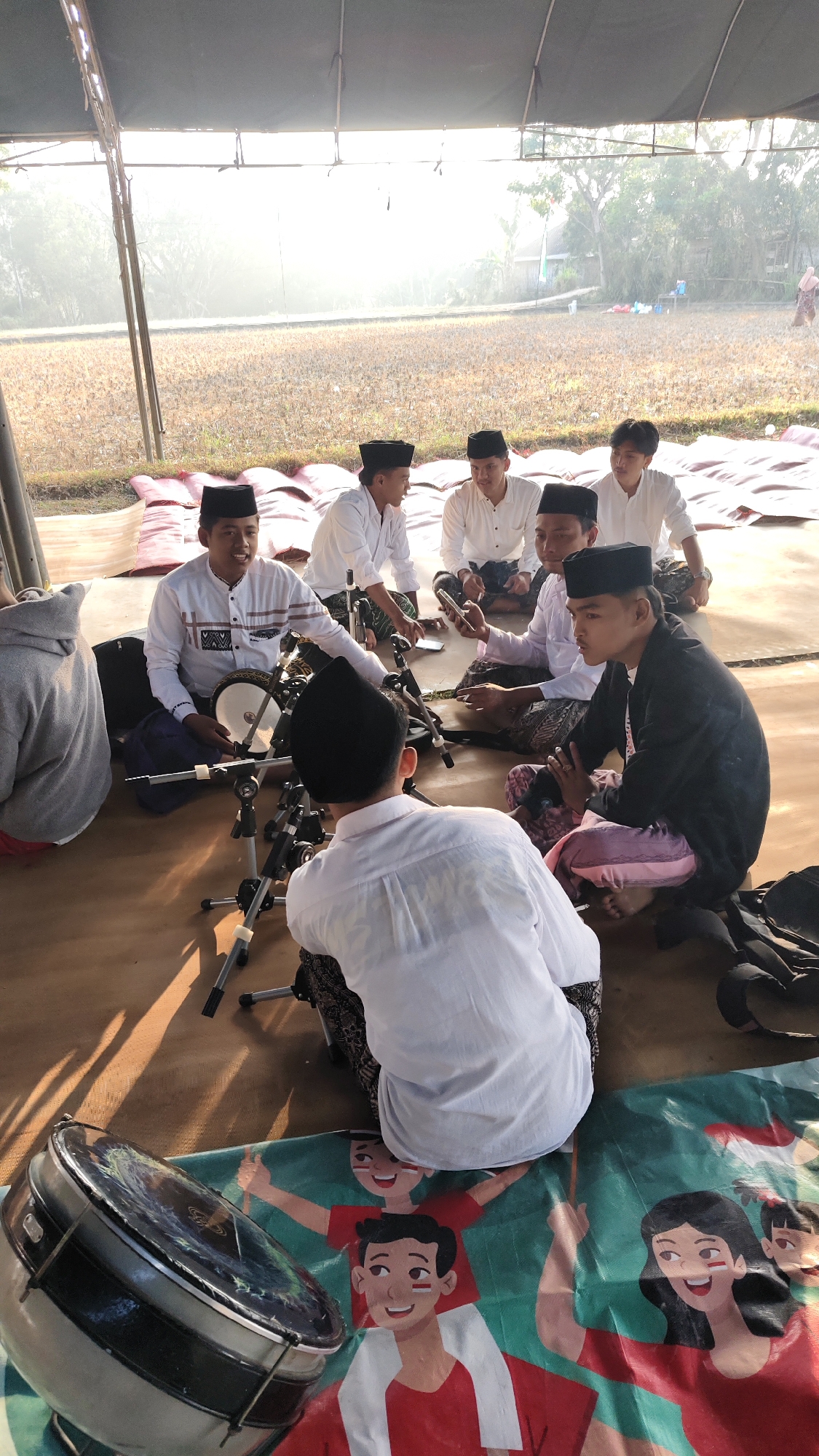 In Galis Daya, Indonesia, under a tent on a dirt-covered ground, a group of young men are gathered. They are seated on thin red mats arranged on the ground. Seven individuals are visible, all wearing white shirts and black peci hats, common religious attire. Two of the men are playing traditional instruments. One man in the foreground is seated facing away from the camera, with his back towards the viewer, looking at a drum on a stand. Another man, slightly to the left, is playing a small drum while looking towards the others. A third man, seated to the right, is looking at his phone. In the background, two more young men are visible, one of whom is also looking at his phone. The environment suggests an outdoor setting, possibly a field, with dry vegetation visible beyond the tent. The lighting indicates it is daytime, with a bright, hazy sun filtering through the tent. The overall scene depicts a gathering of young men engaged in musical practice or a performance.