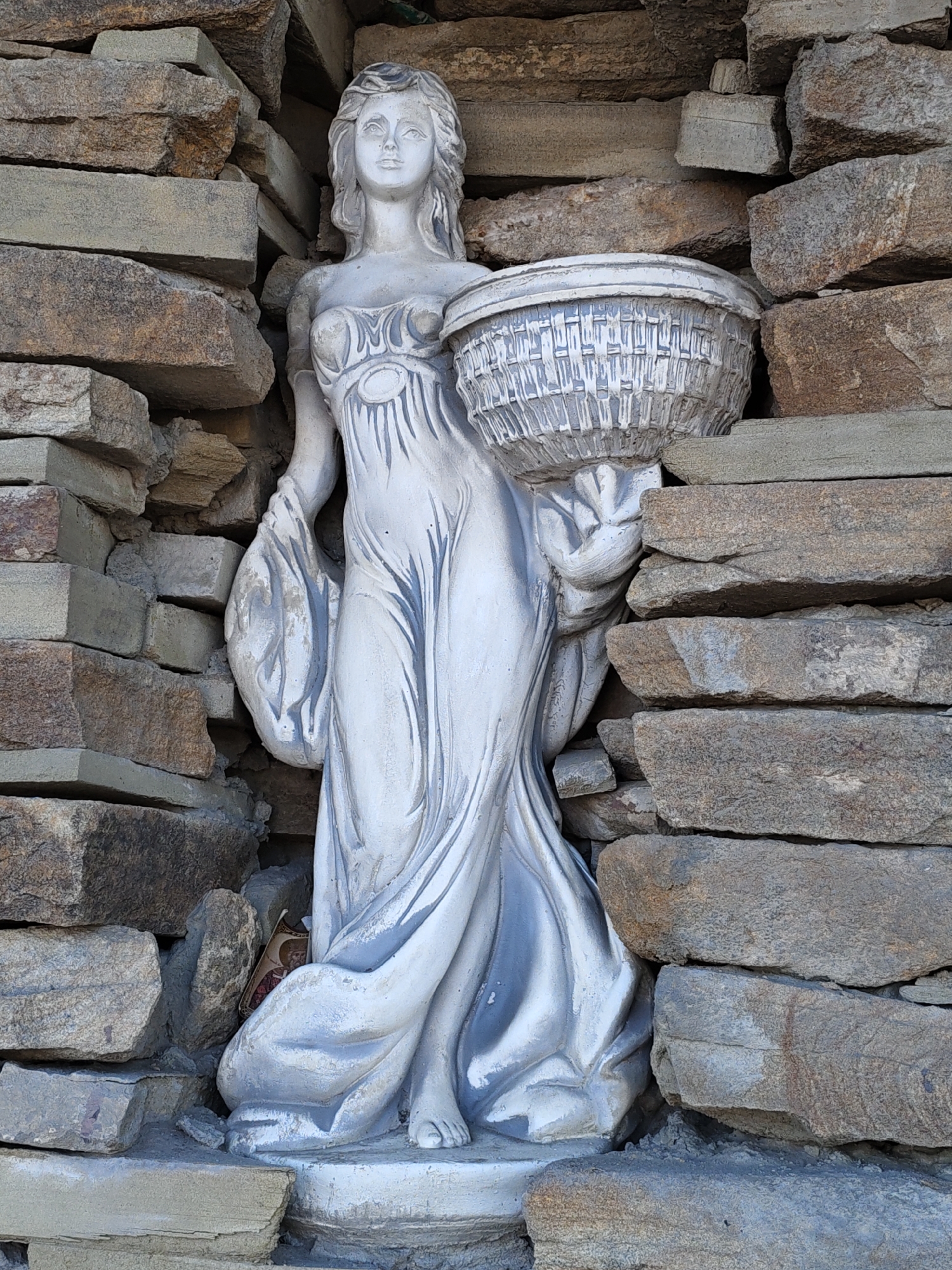 The image shows a weathered, pale statue of a woman holding a basket, nestled within a wall constructed of rough, stacked stones. The statue is light gray and appears to be made of concrete or plaster, showing some signs of wear and tear, with chipped edges and faded paint. The woman is depicted in a flowing robe, with her hair styled in waves. She is holding a woven basket in her right arm, and a bundle of fabric or cloth is draped over her left arm. The stone wall surrounding the statue is composed of various sizes and colors of natural stone, primarily earthy tones of brown, gray, and tan, with some lighter gray stone blocks forming the base. The stones are stacked unevenly, creating a rustic and somewhat aged appearance. A small, colorful religious icon or picture is partially visible tucked into the fold of the statue's robe near its feet. The overall environment suggests an outdoor setting, possibly a garden or courtyard, with the textured stone and weathered statue contributing to a sense of age and perhaps neglect. The statue is centrally located within the frame, with the stone wall acting as a backdrop and frame for the subject. The lighting suggests it is daytime, with shadows cast by the stones and the statue. The scene is static, with no discernible activity or interaction occurring.