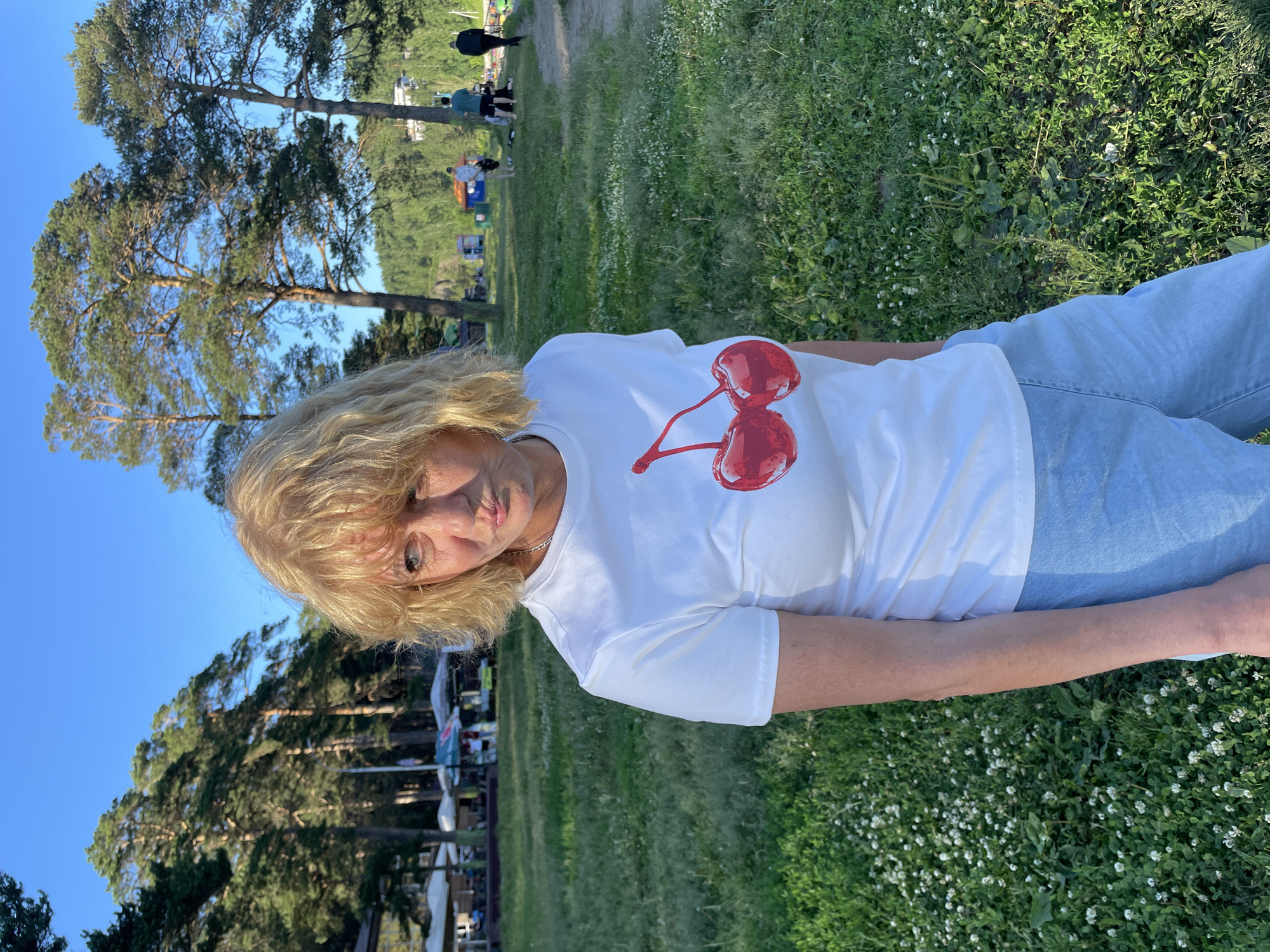 The image shows a medium shot of a woman in her 60s, with blonde, wavy hair, looking towards the camera. She is wearing a white t-shirt with a graphic of two red cherries and light blue denim jeans. The woman is positioned in the foreground, with the background showing a grassy area with scattered white flowers and trees. In the distance, there are more trees and some people, suggesting an outdoor gathering or park setting. The lighting indicates it's daytime, likely late afternoon due to the warm, soft light. The overall mood appears relaxed and casual. The image is oriented vertically.