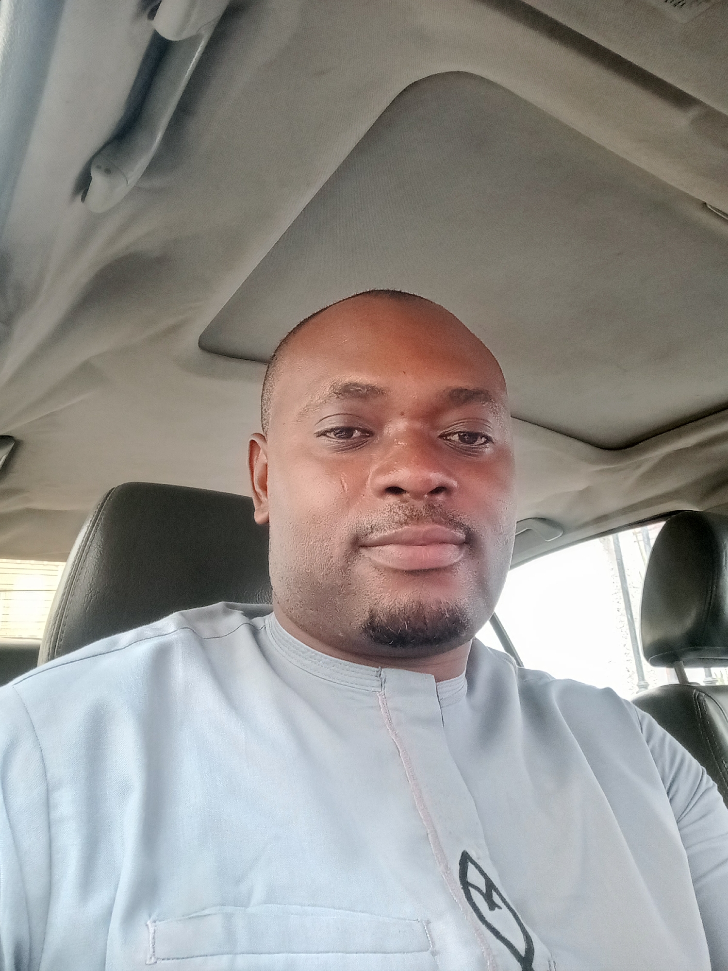 A medium shot depicts a dark-skinned man with a shaved head, seated in a car. He is wearing a light blue traditional garment with a dark design on the front. The car's interior is visible, including the ceiling with a sunroof, a rear-view mirror, and the driver's seat. The location is Gwarinpa, Nigeria, though no specific landmarks are visible. The man is looking directly at the camera.