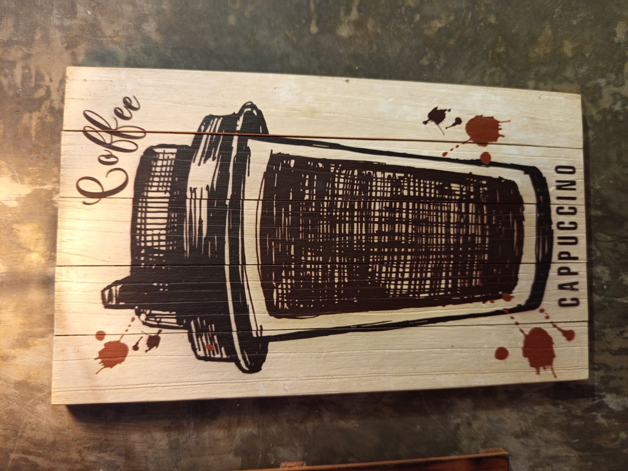 A slightly angled, medium shot shows a coffee-themed wooden sign. The sign is rectangular and made of horizontal wooden planks painted in an off-white color. A dark brown drawing of a coffee cup with the words "Coffee" (vertically on the left) and "Cappuccino" (vertically on the right) is on the sign. The image is in a building in North Kuta, Indonesia. The sign also has decorative brown paint splatters. The surface under the sign appears to be a textured wall with shades of gray.