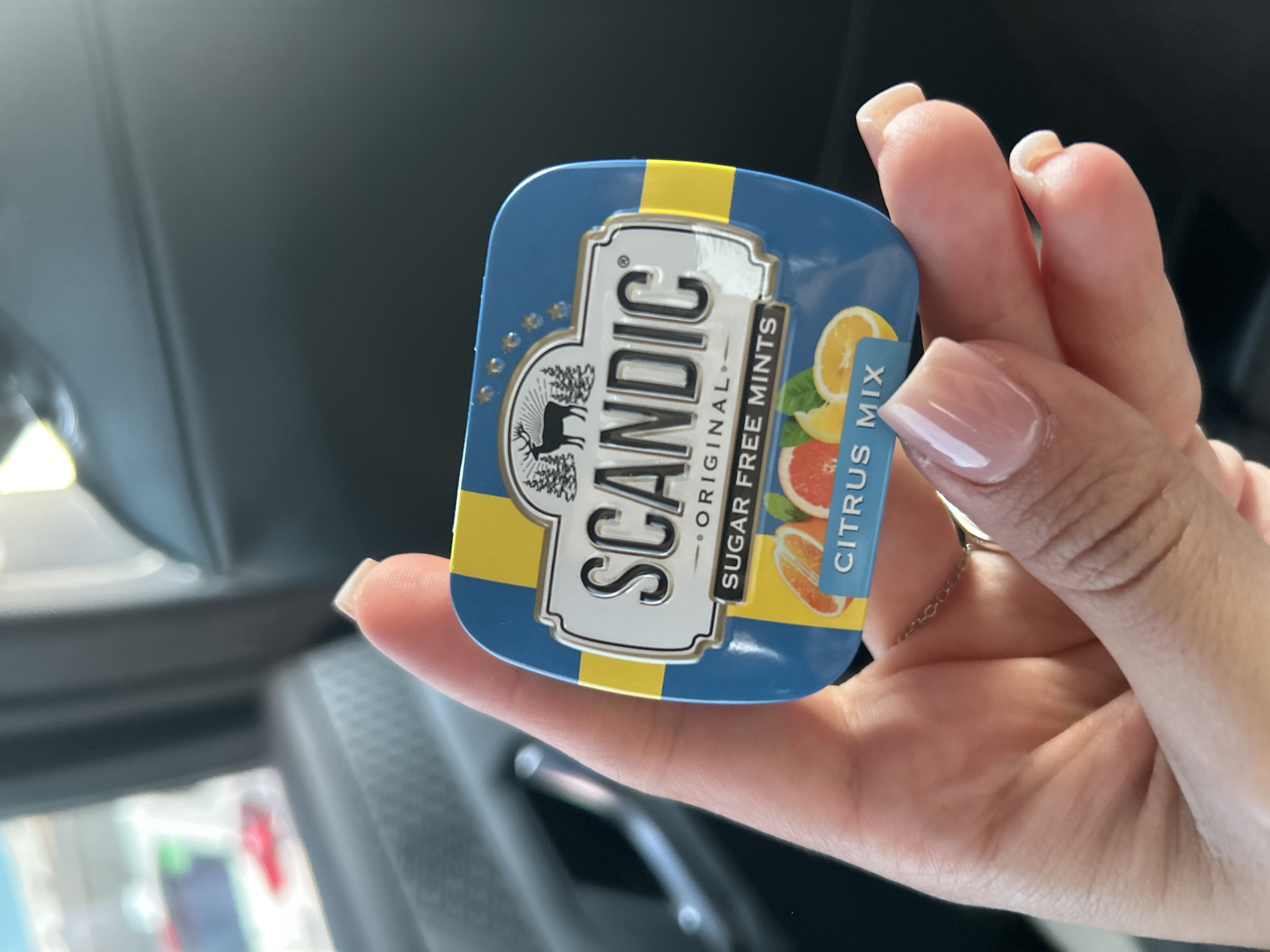A close-up, top-down shot shows a person's hand holding a small, rectangular tin of Scandic Original Sugar Free Mints in Citrus Mix flavor. The tin has a blue and yellow striped design, with a white label featuring the Scandic logo, a silhouette of a deer and trees, and images of citrus fruits. The background is out of focus and appears to be the interior of a car, with a dark seat and a glimpse of a window with cars outside. The lighting suggests it is daytime.